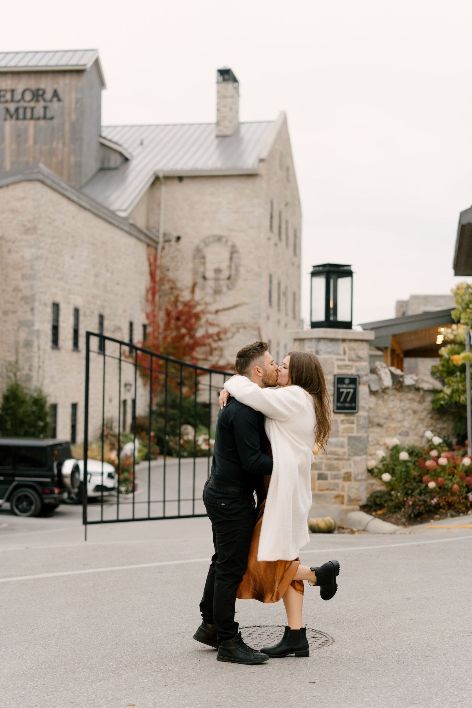 Couple sharing a romantic kiss in front of the Elora Mill entrance gate during an autumn engagement session.