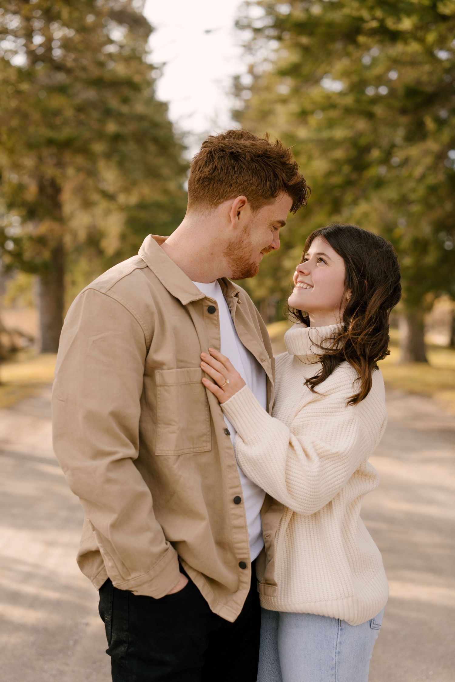 Smiling couple embracing on a tree-lined path, woman in cream sweater and jeans looks up lovingly.