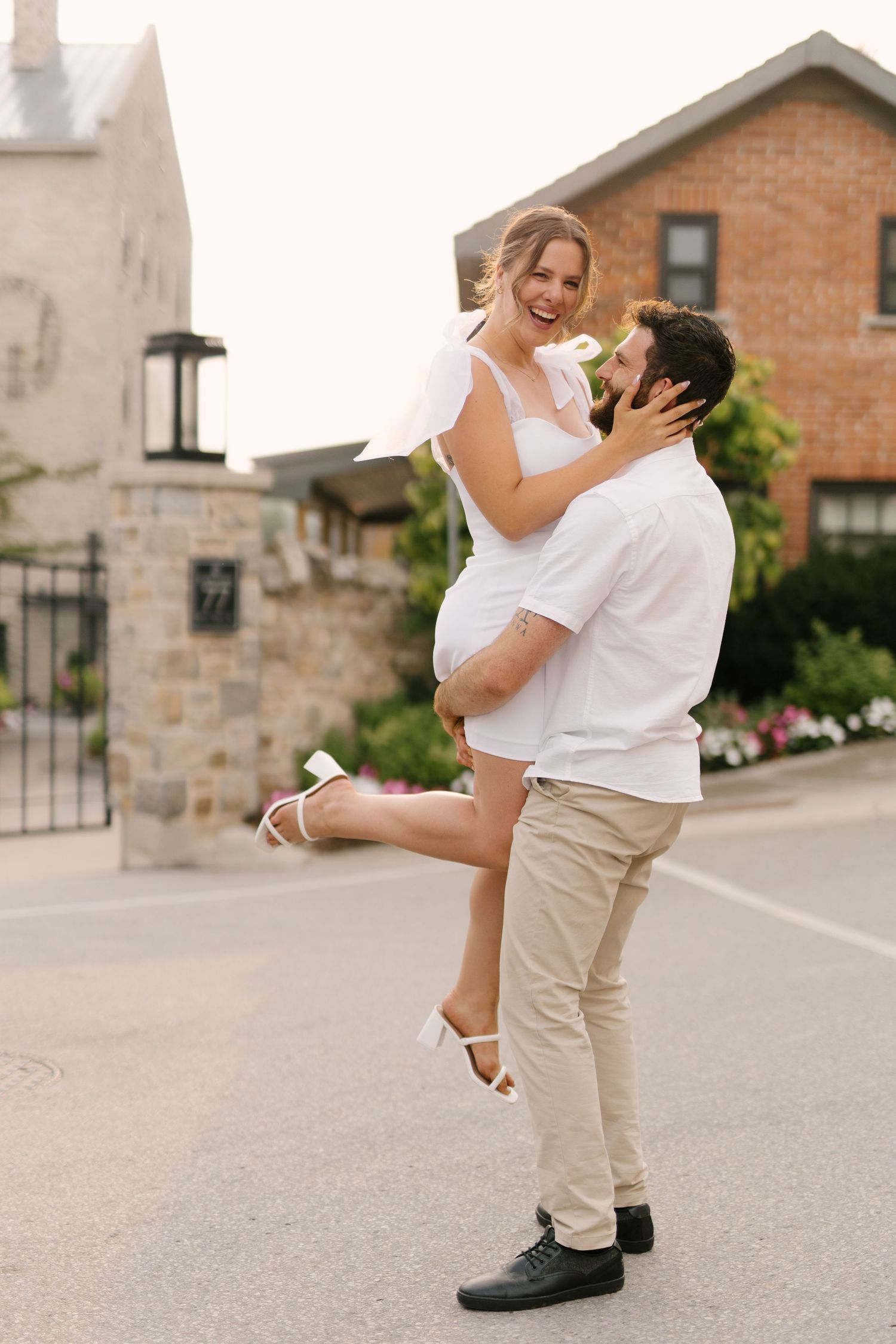 Bearded man lifting and spinning his laughing fiancee in white dress on a sunny street.