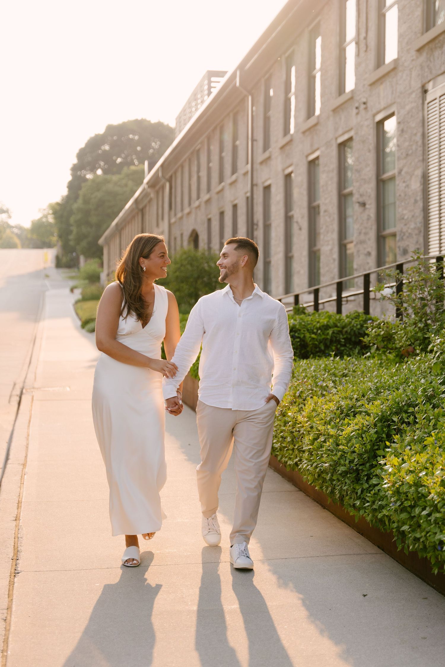 A happy couple in white outfits strolls hand-in-hand along a sunlit path beside a stone building.
