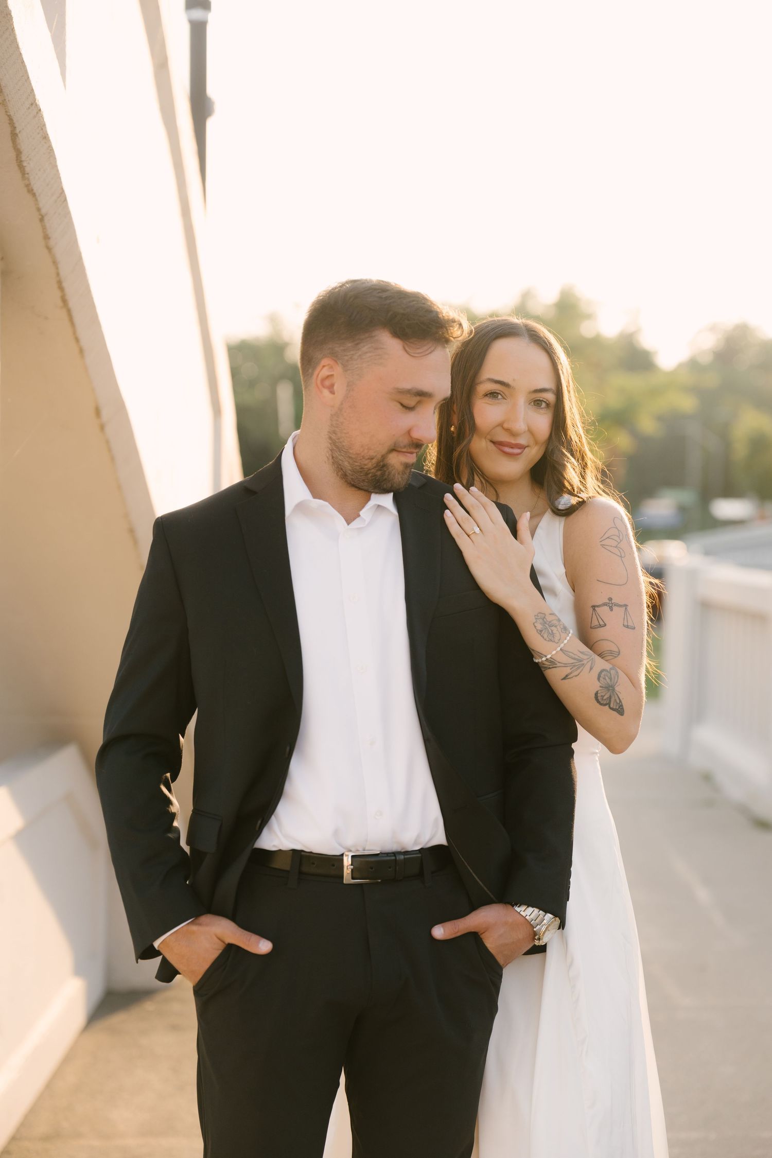 Engaged couple posing on bridge at sunset, woman leaning on mans shoulder showing ring.