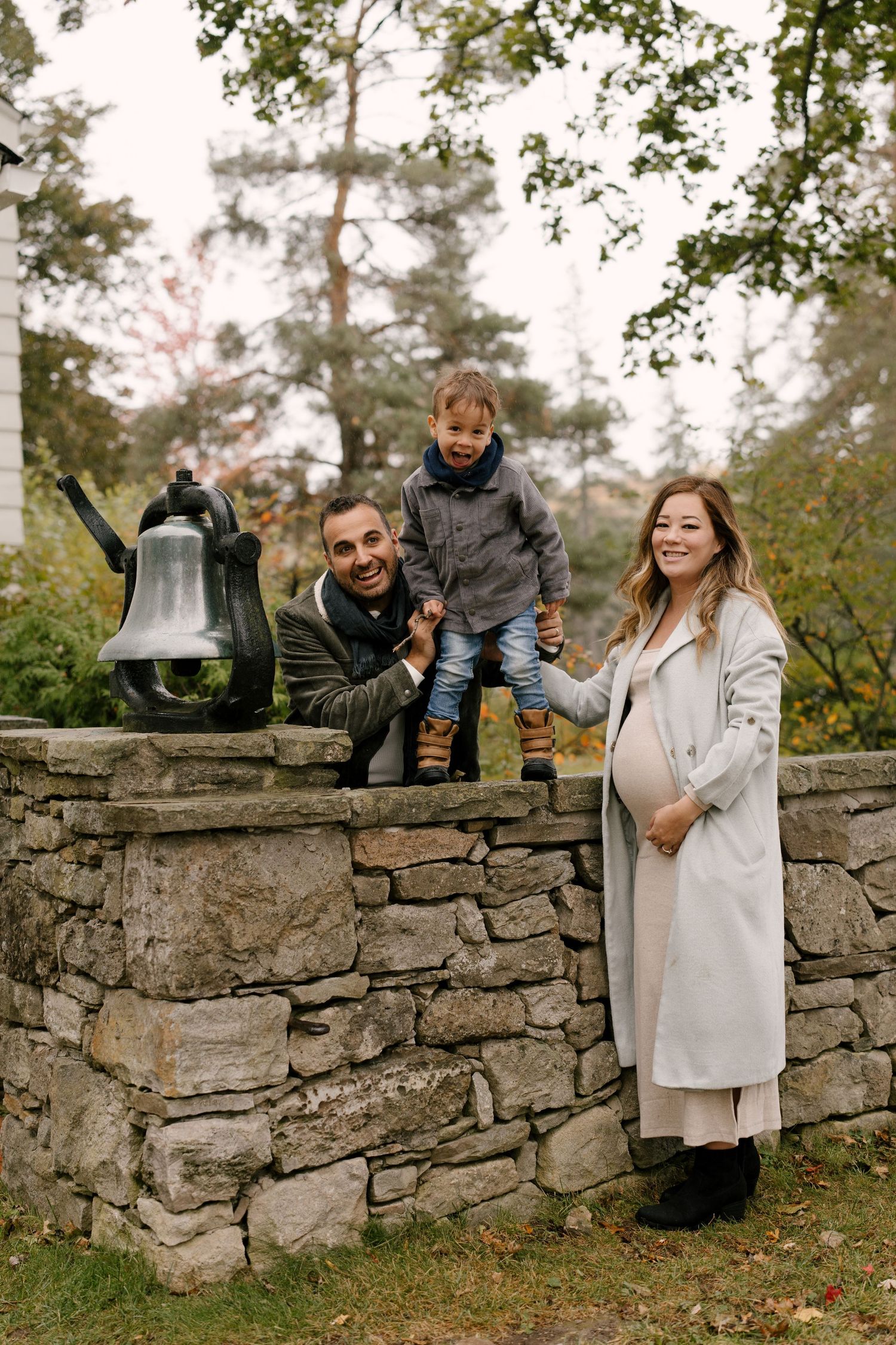 Smiling family with pregnant mother posing by stone wall with bell in autumn park setting.
