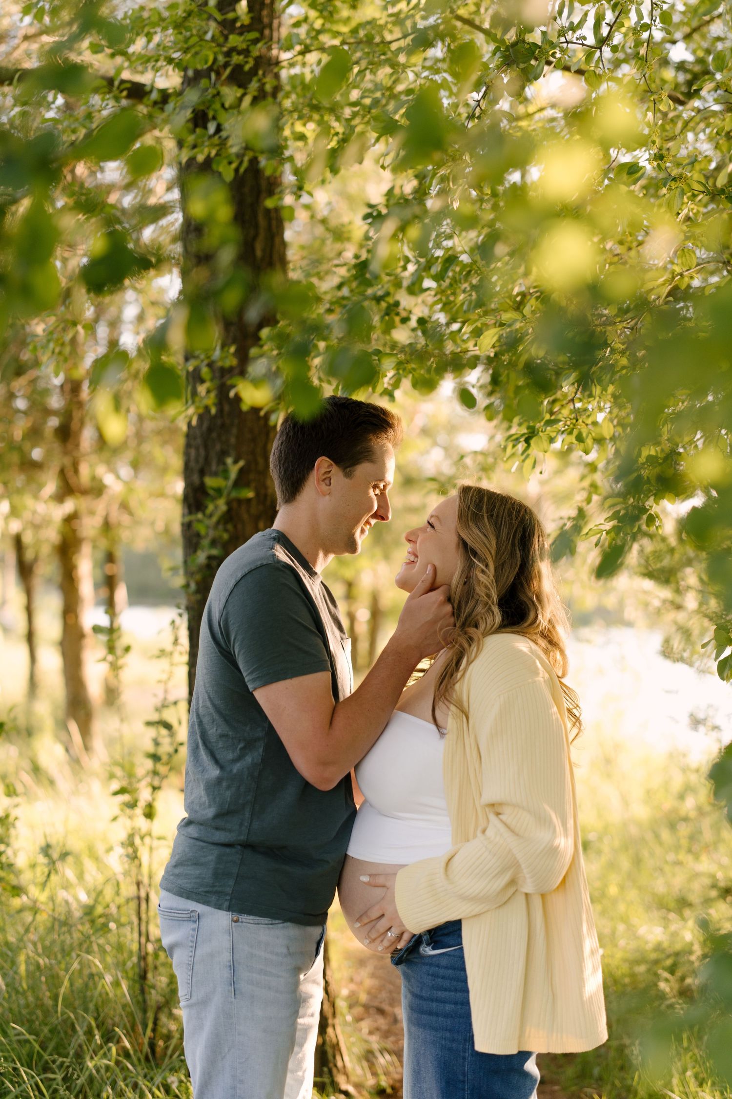 Couple shares a romantic moment under leafy trees, man gently holds woman's face near a sunlit lake.