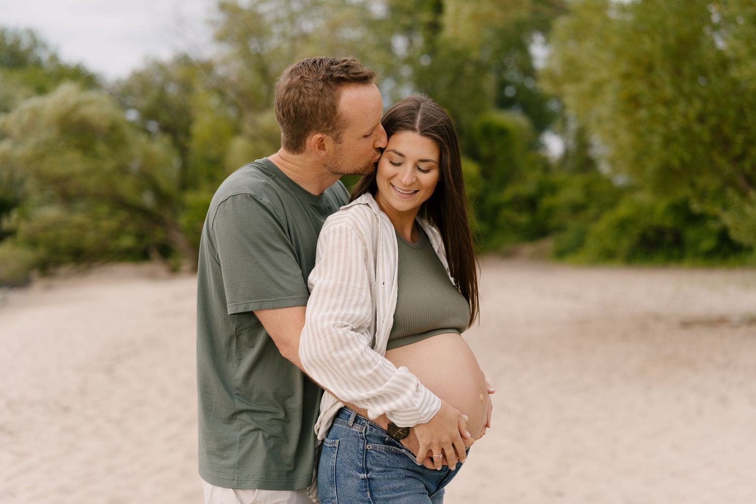 Happy couple outdoors, man kissing pregnant womans cheek while embracing her baby bump on sandy beach.