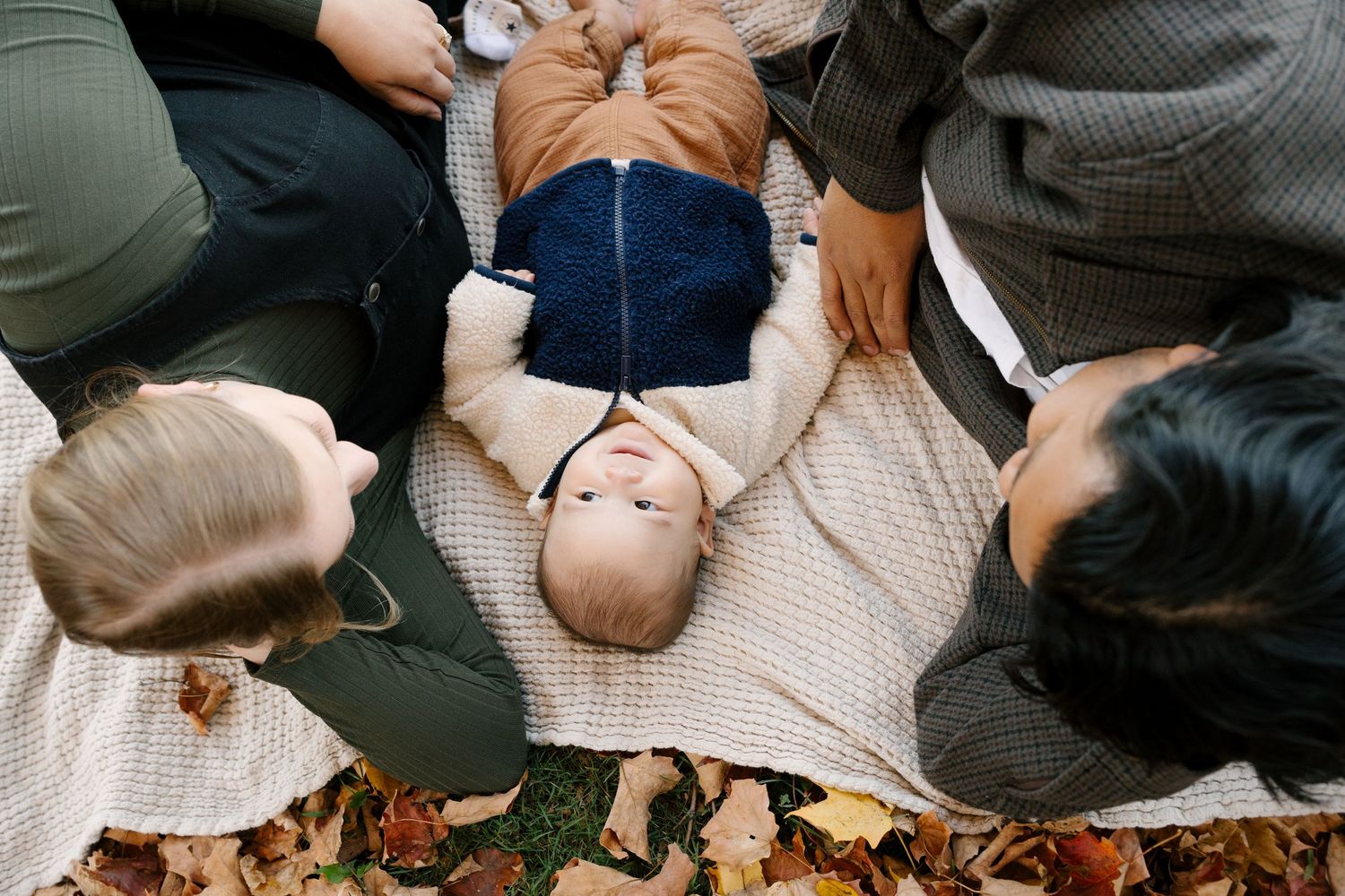 Overhead view of baby lying on blanket surrounded by parents in autumn leaves.