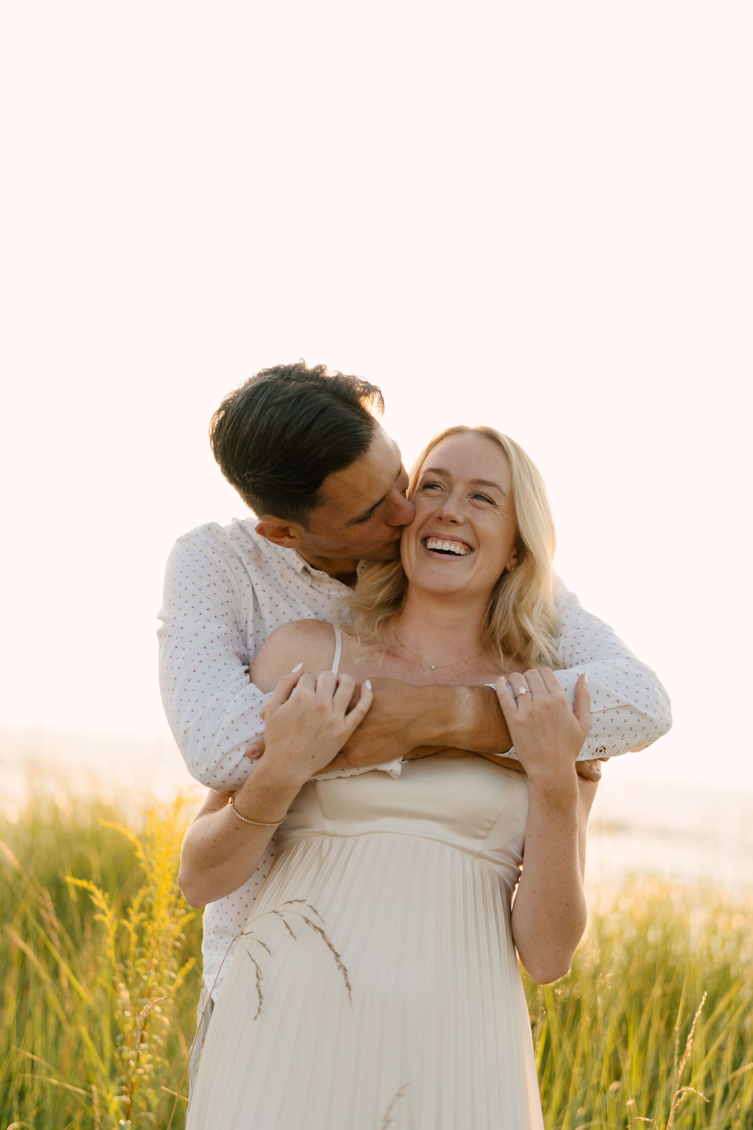Man kissing laughing woman in golden grass field at sunset.