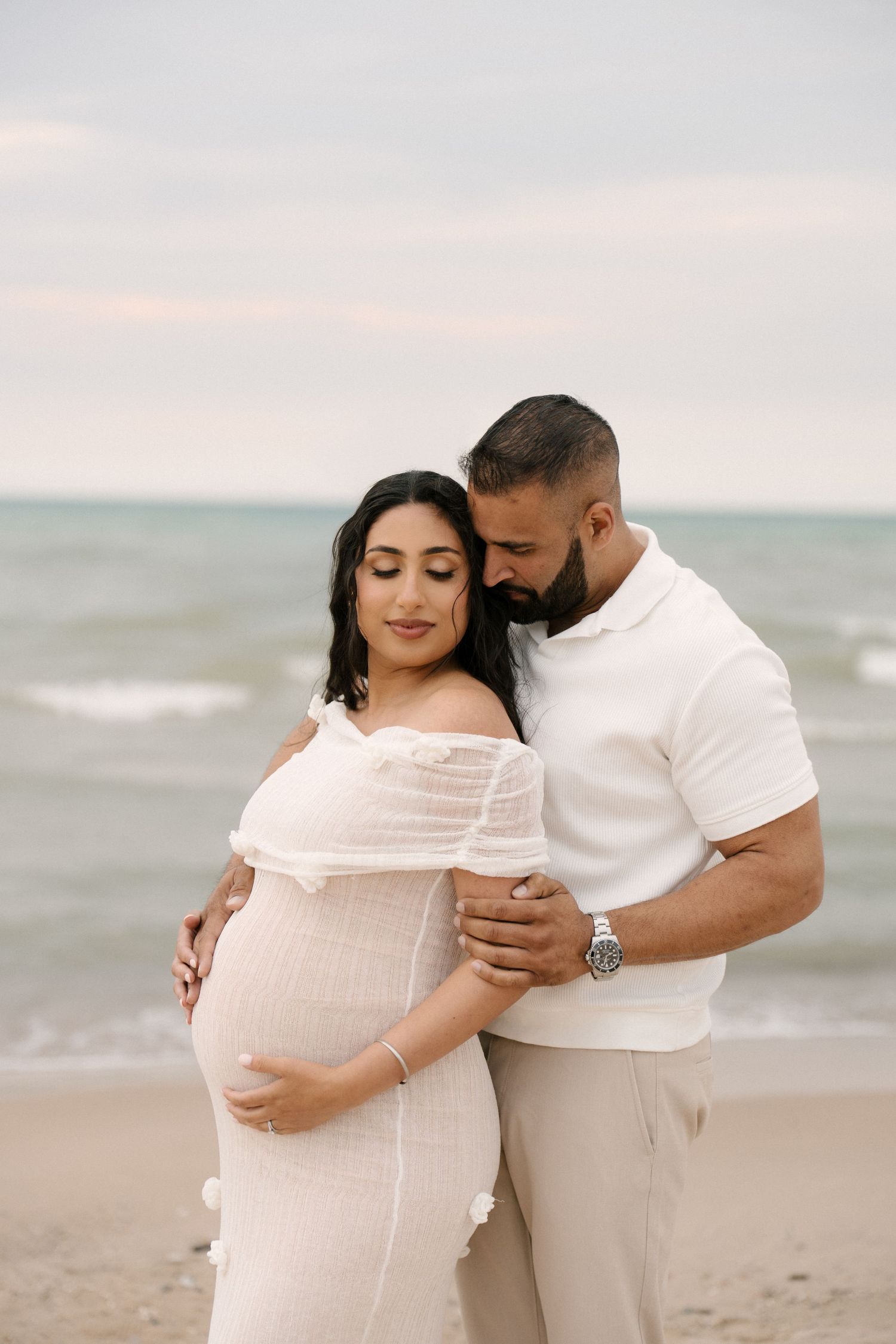 Couple in white outfits sharing tender maternity beach portrait at sunset by the ocean.