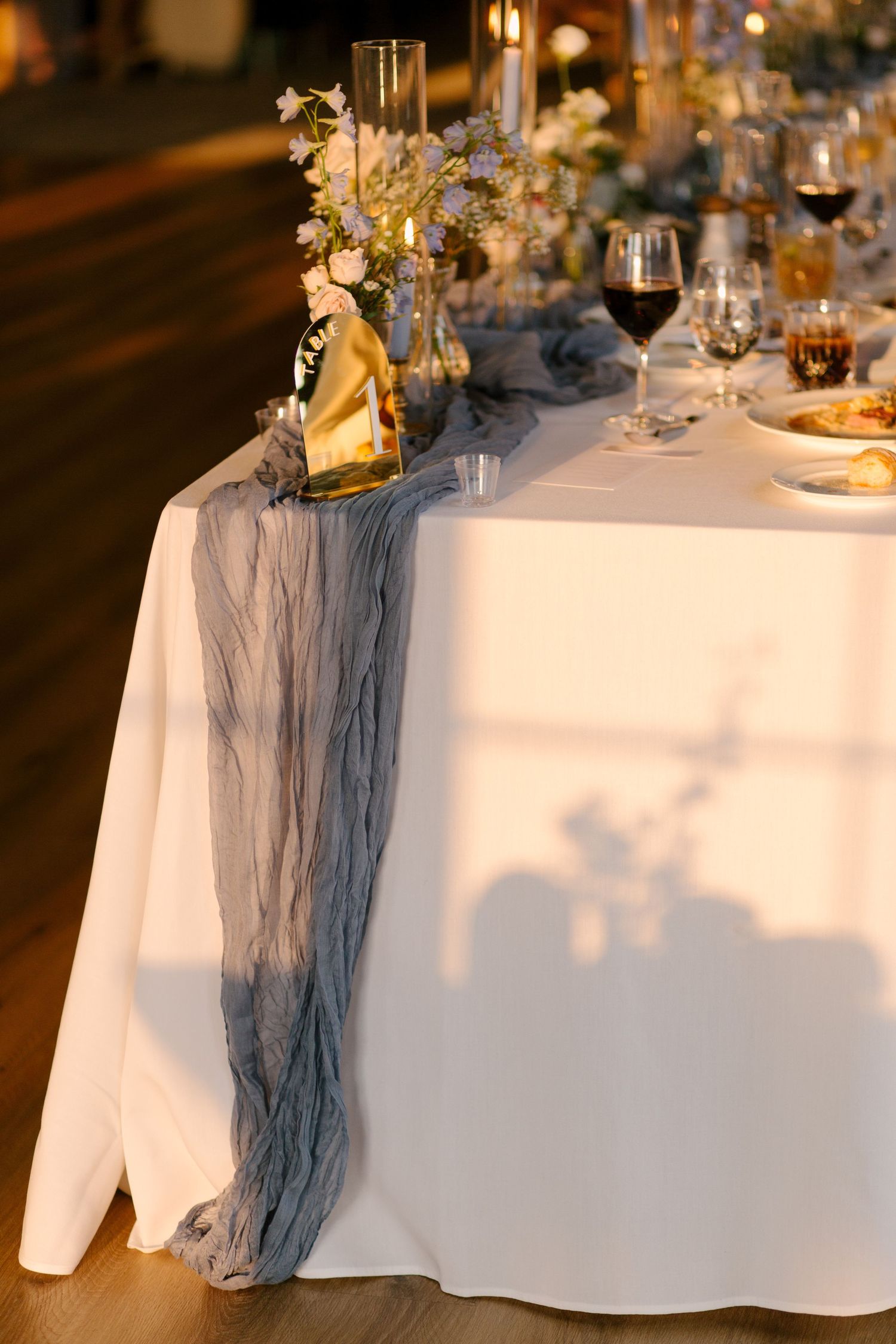 Elegant wedding reception table with dusty blue gauze runner, candles, and floral centerpieces at night.