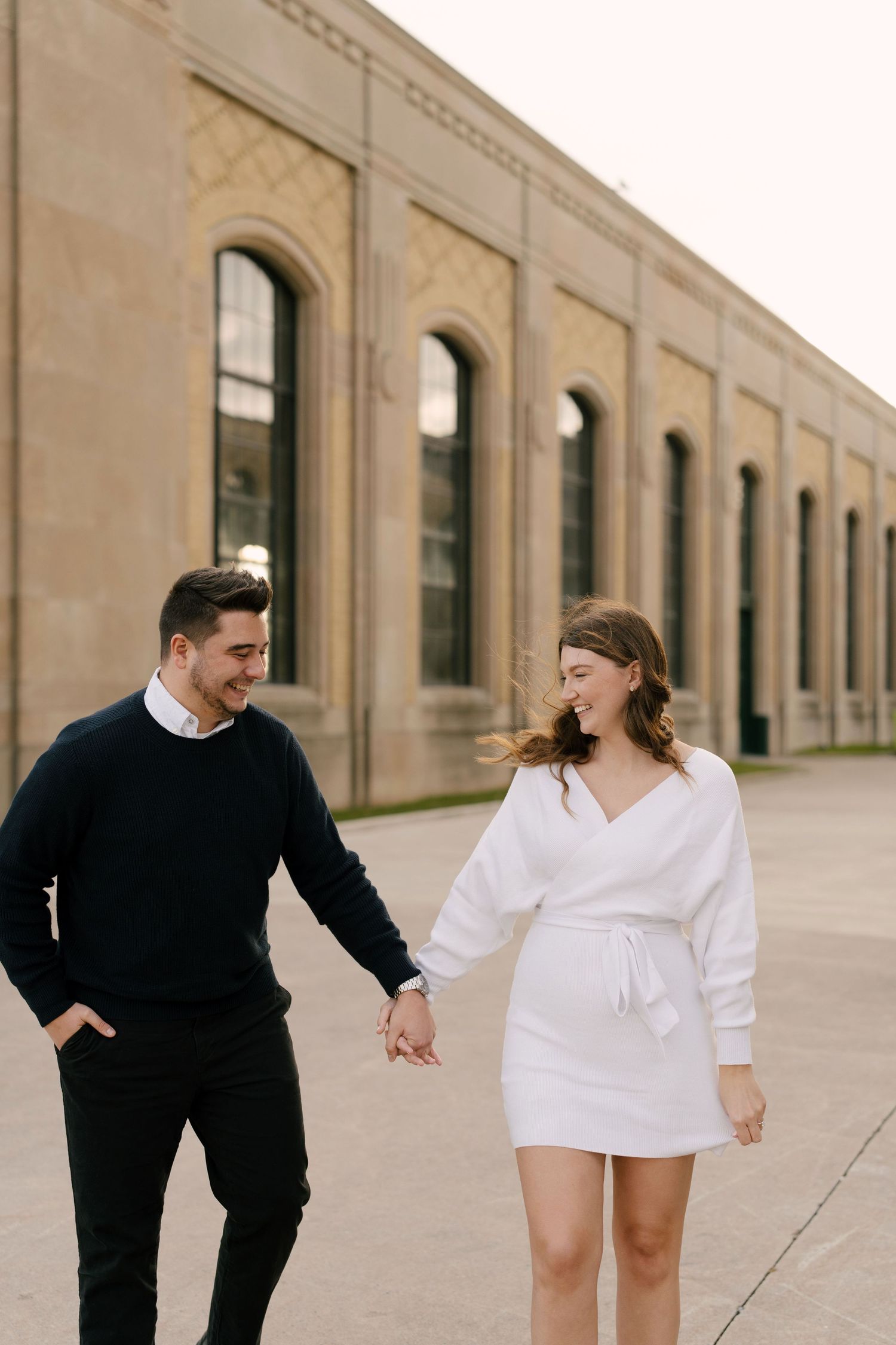 Smiling couple holding hands and walking together past a large stone building outdoors.