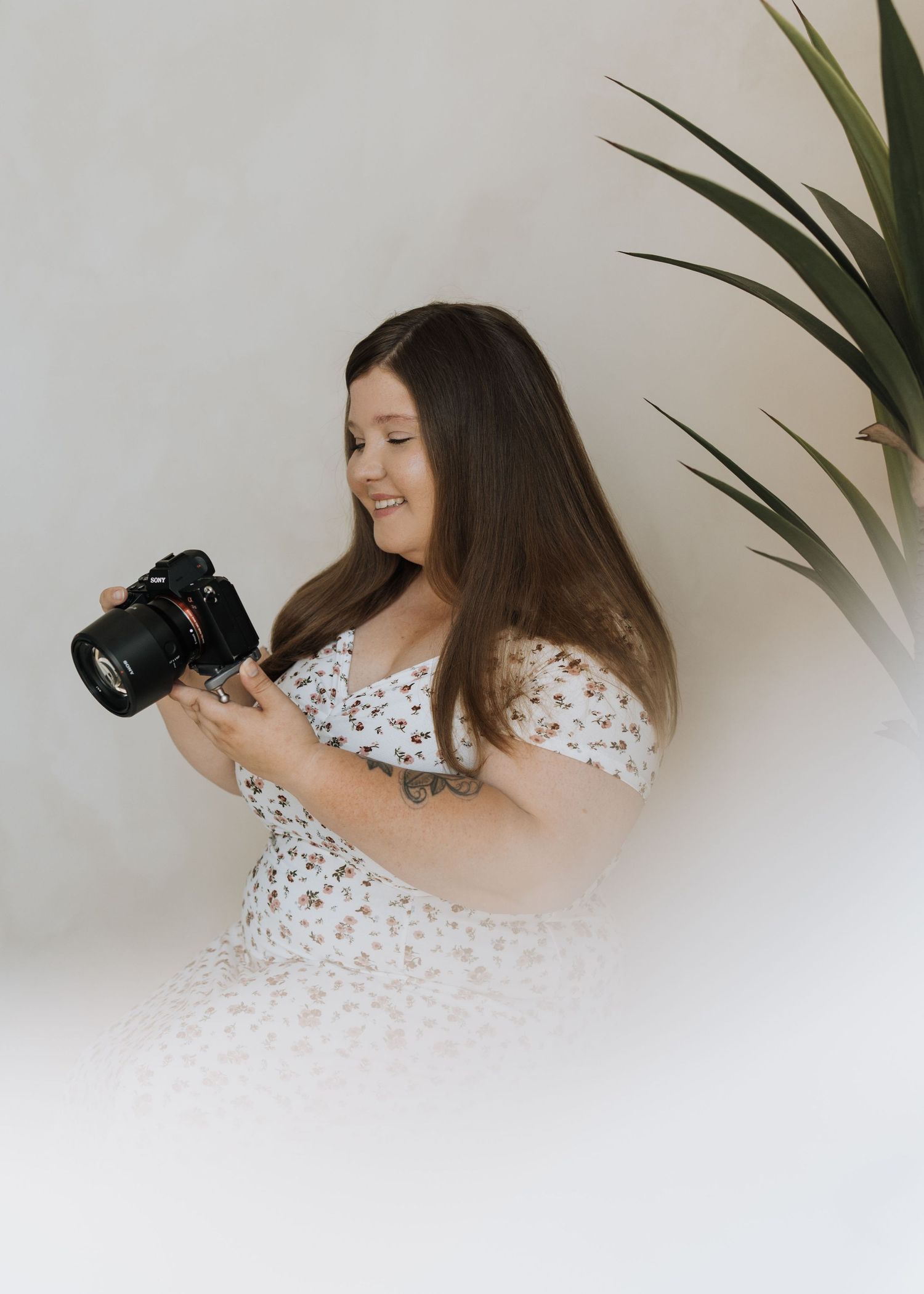 Smiling female photographer holding DSLR camera indoors beside tropical plant on white background.