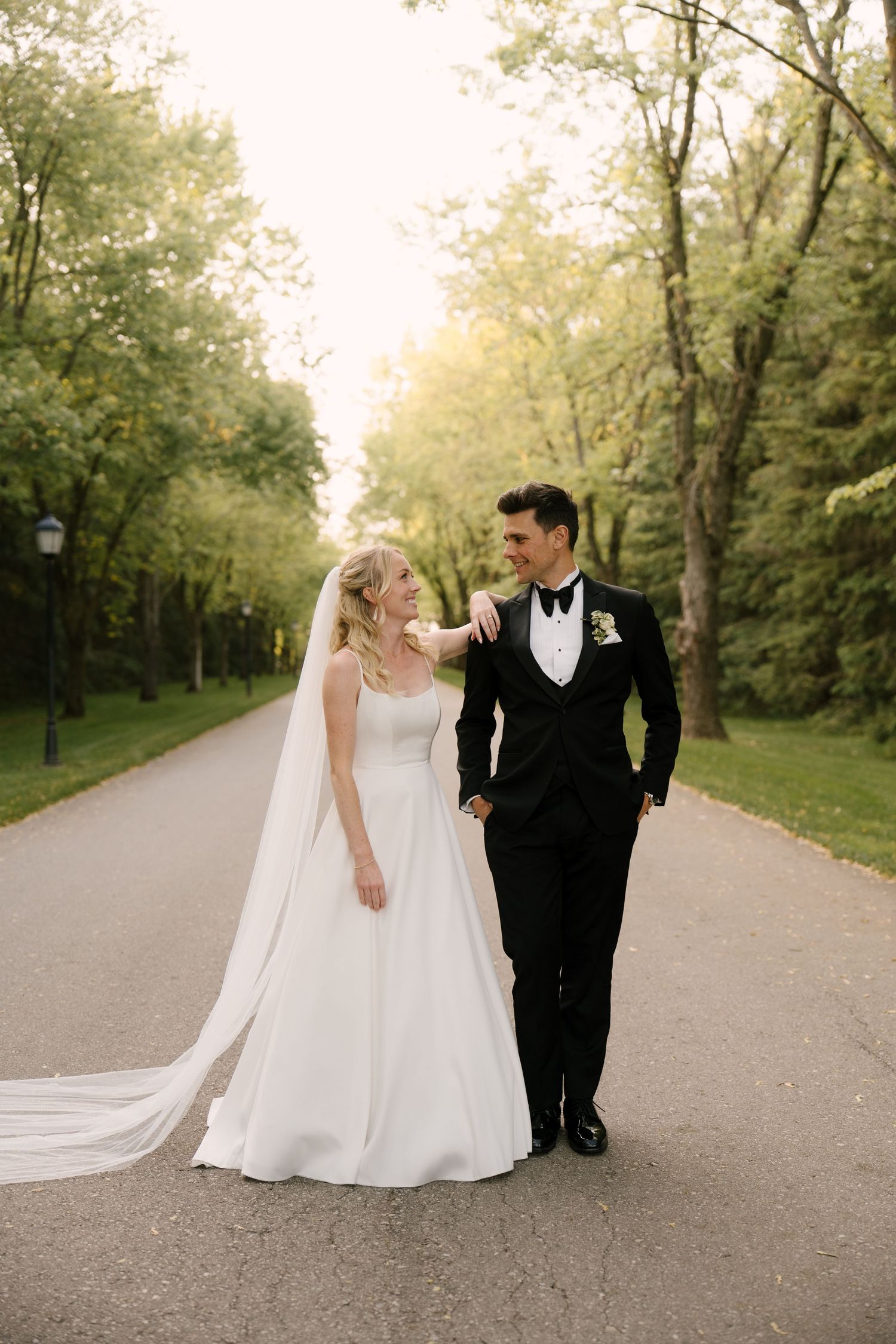 Bride and groom walking hand in hand down a tree-lined path in golden hour light at Roseville Estates wedding venue.