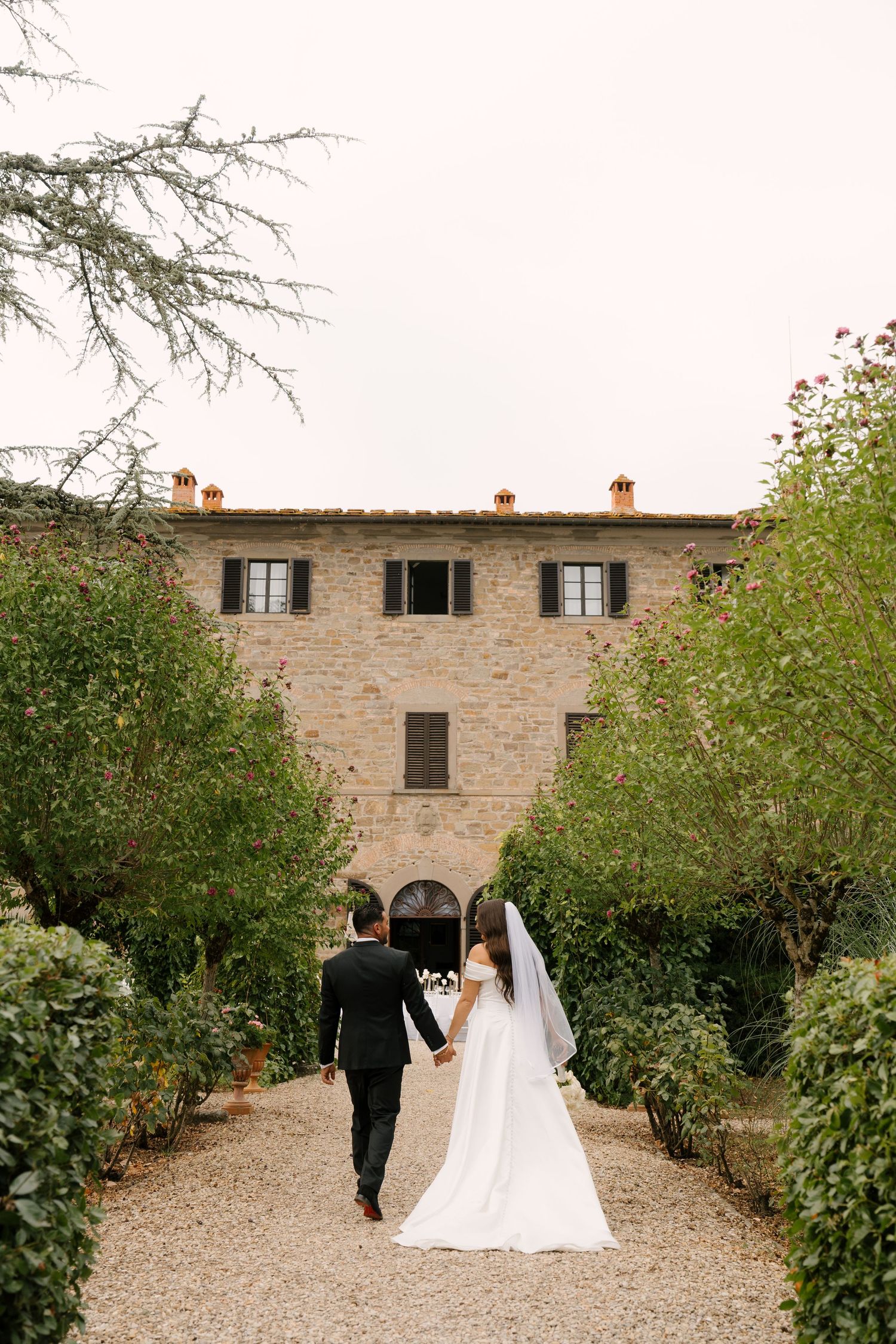 Newlywed couple walks hand-in-hand toward rustic Tuscan stone villa surrounded by lush greenery on gravel path.