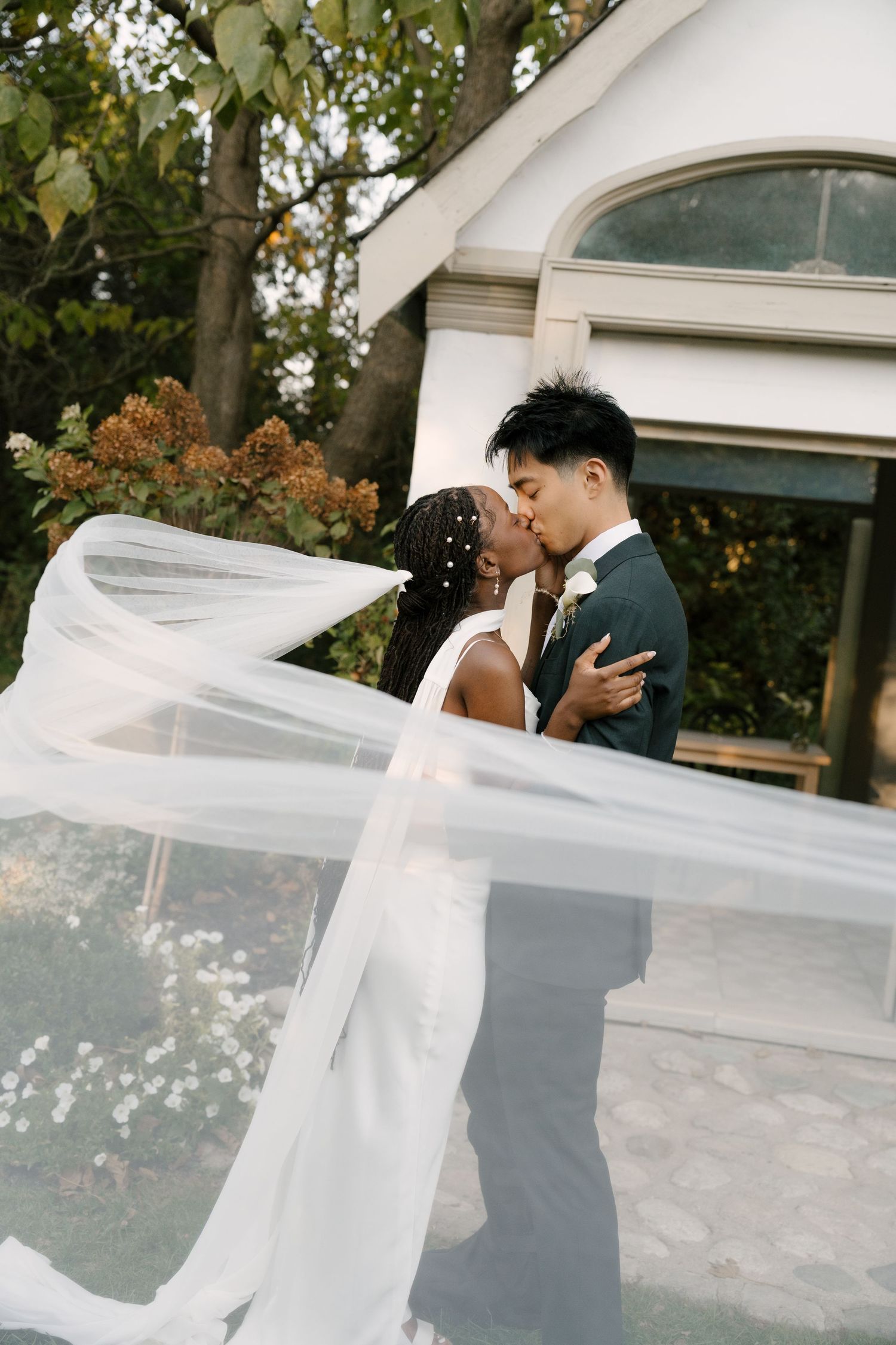 An interracial couple shares a kiss with the bride's veil flowing dramatically in the wind outside a white chapel at Wren House.