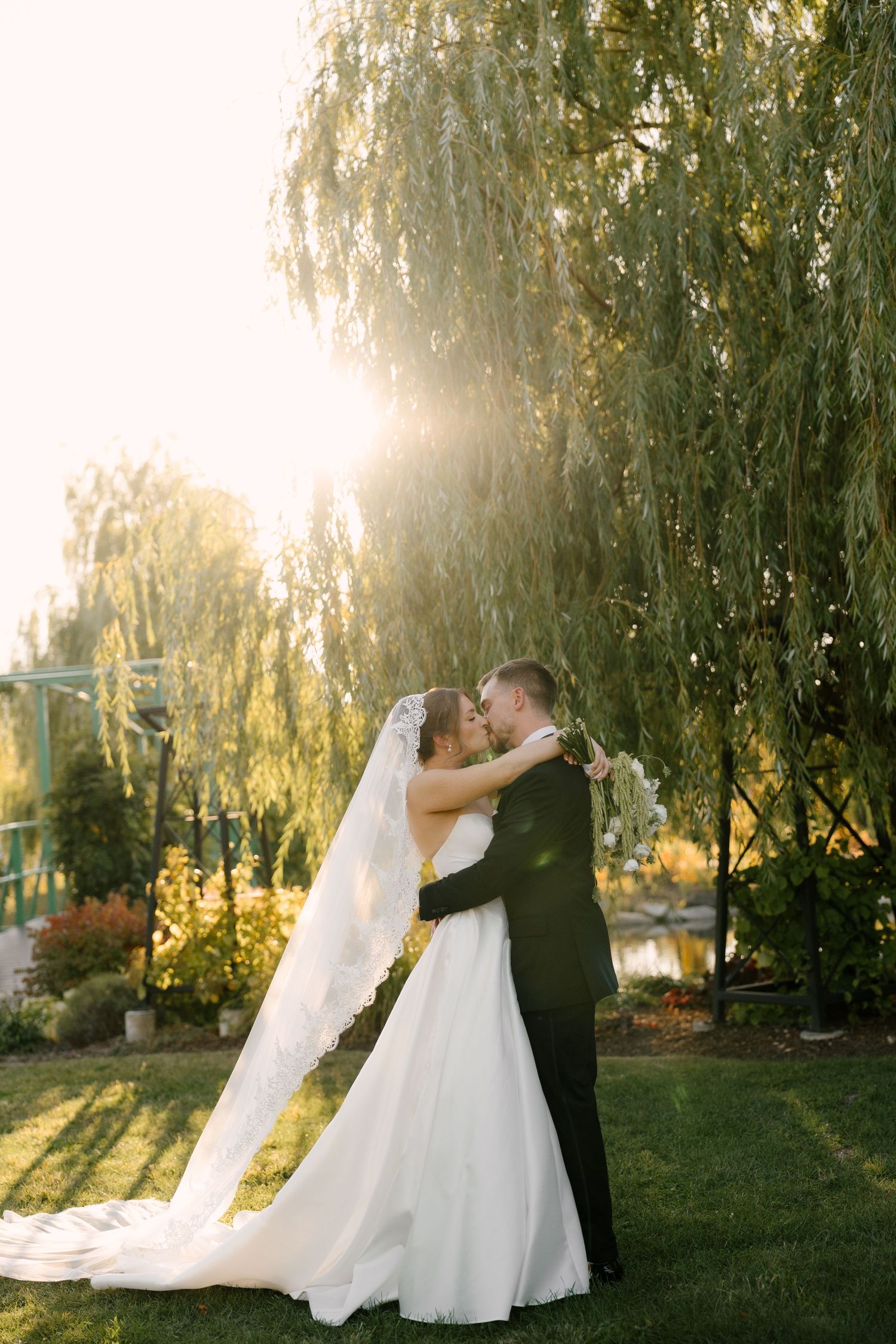 Bride and groom kissing under a weeping willow tree at golden hour in a lush garden setting at Pillar and Post wedding venue.