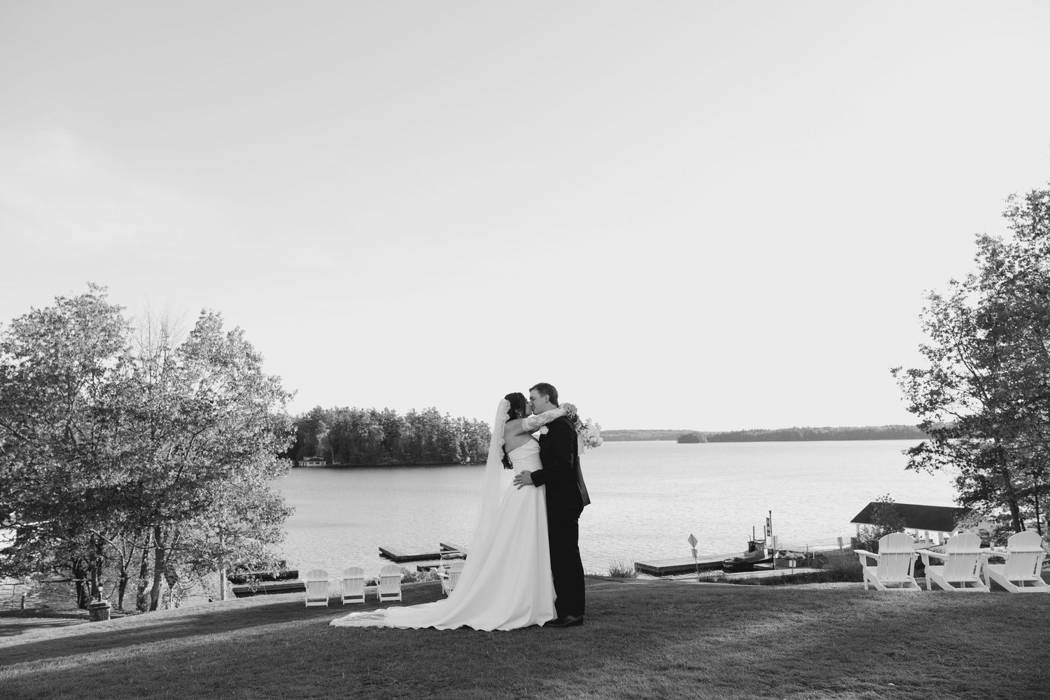 Bride and groom embrace near lakeside dock surrounded by autumn trees, black and white photo.