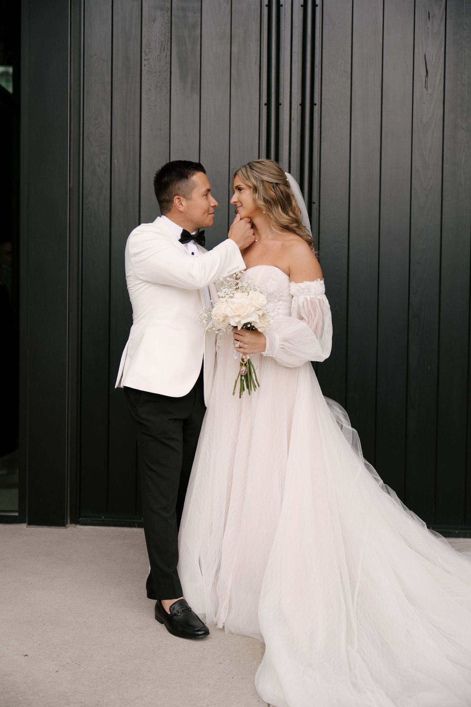Groom in white tuxedo tenderly touches bride's face as they gaze at each other outdoors.