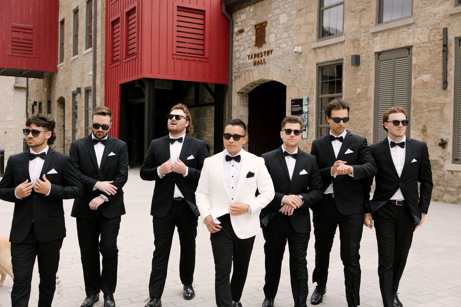 Groom in white tuxedo walks with groomsmen in black suits and sunglasses at outdoor wedding venue.