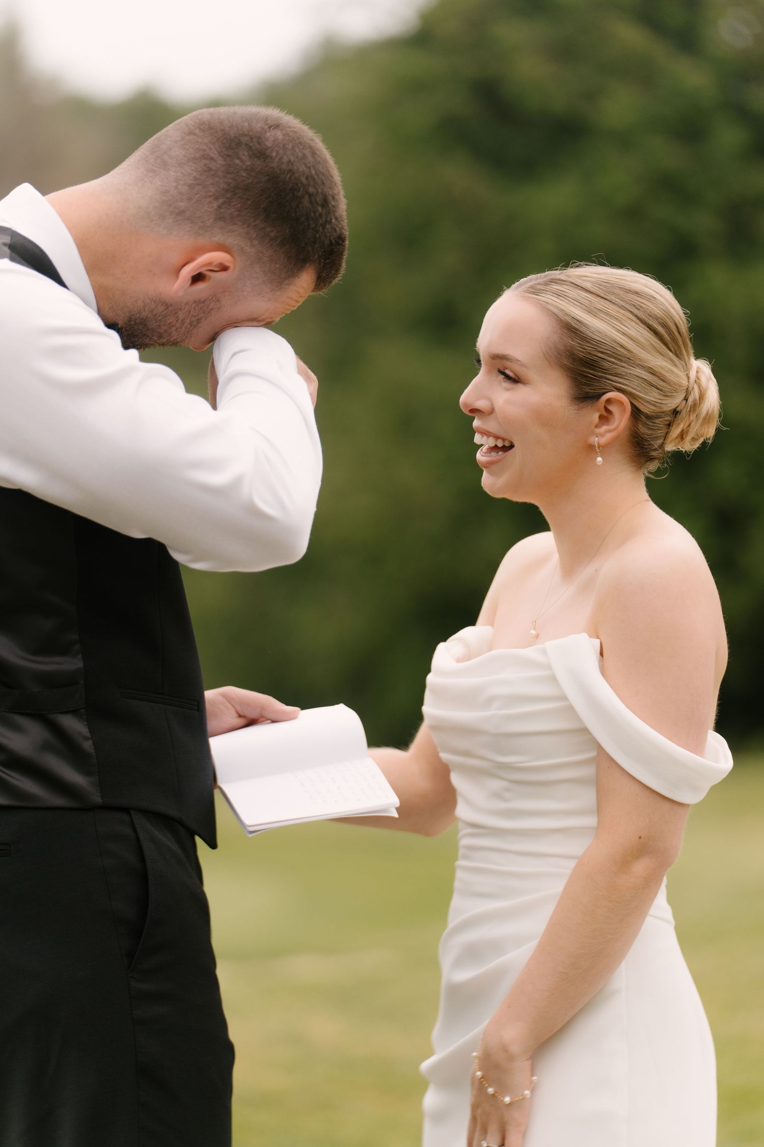 Groom emotionally reading wedding vows from notebook while bride smiles joyfully outdoors.