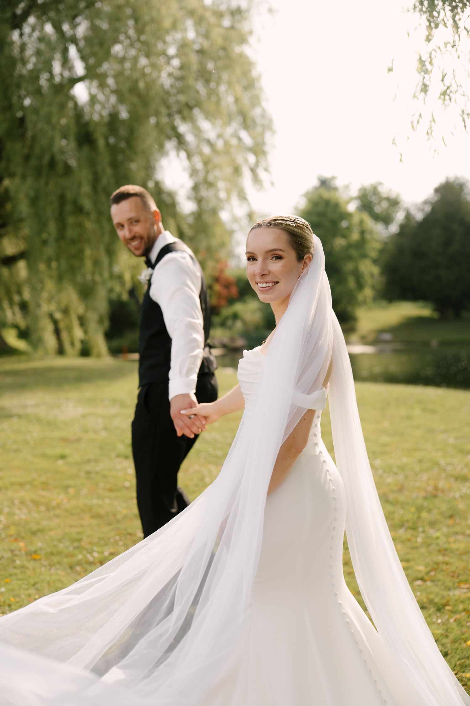Smiling bride in a flowing veil holds grooms hand while walking through a lush green park in Guelph.