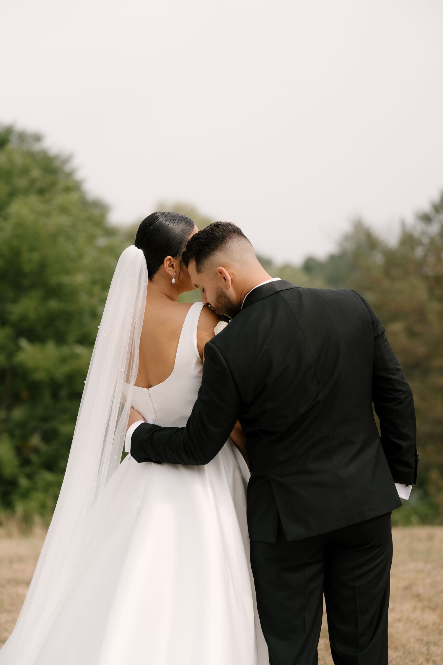 Bride and groom share intimate moment facing away, heads together in open field with trees behind them.