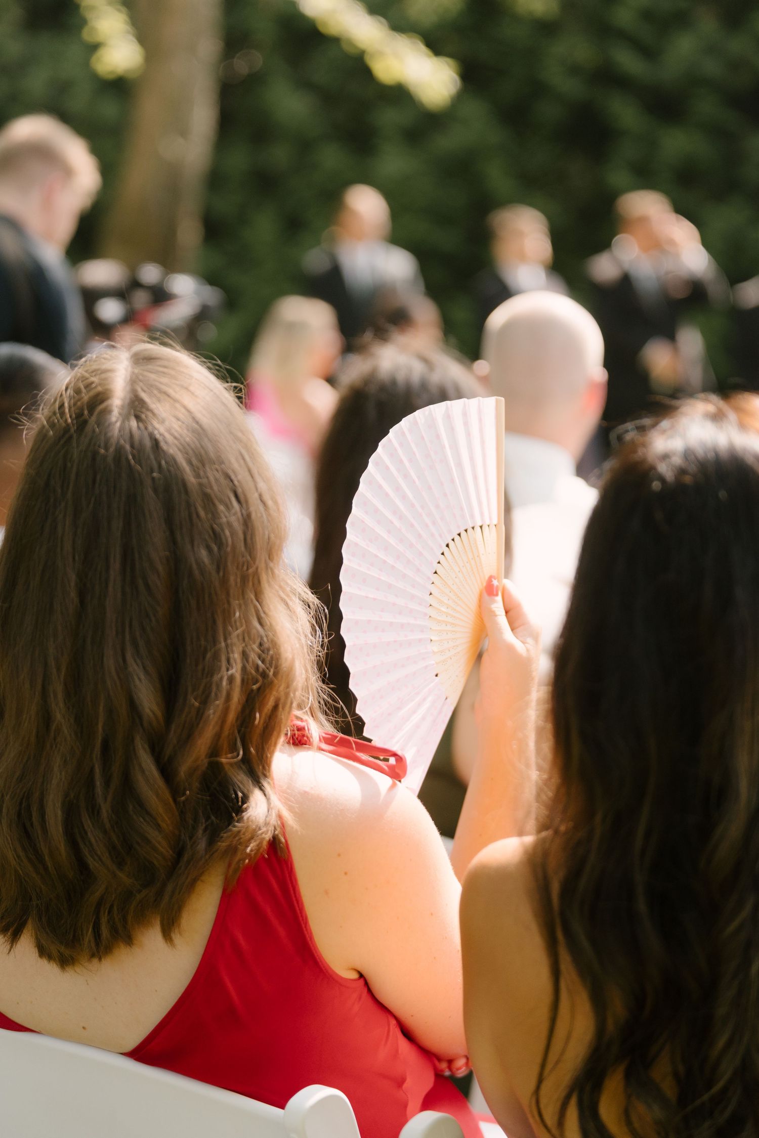 Wedding guests seated outdoors, woman in red dress holding white paper fan on a sunny day.