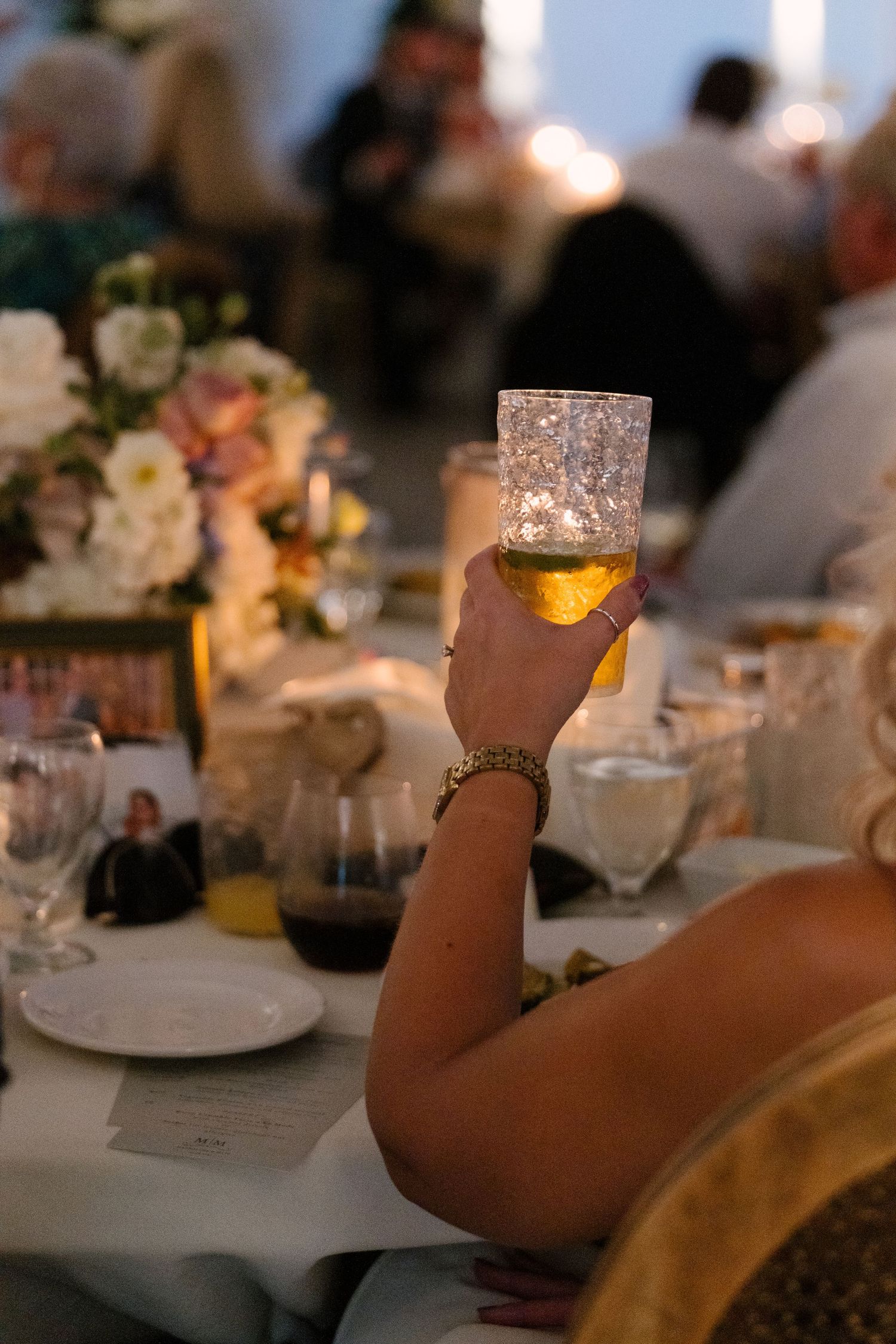 Wedding guest raising a glowing crystal glass during a toast at candlelit reception dinner.