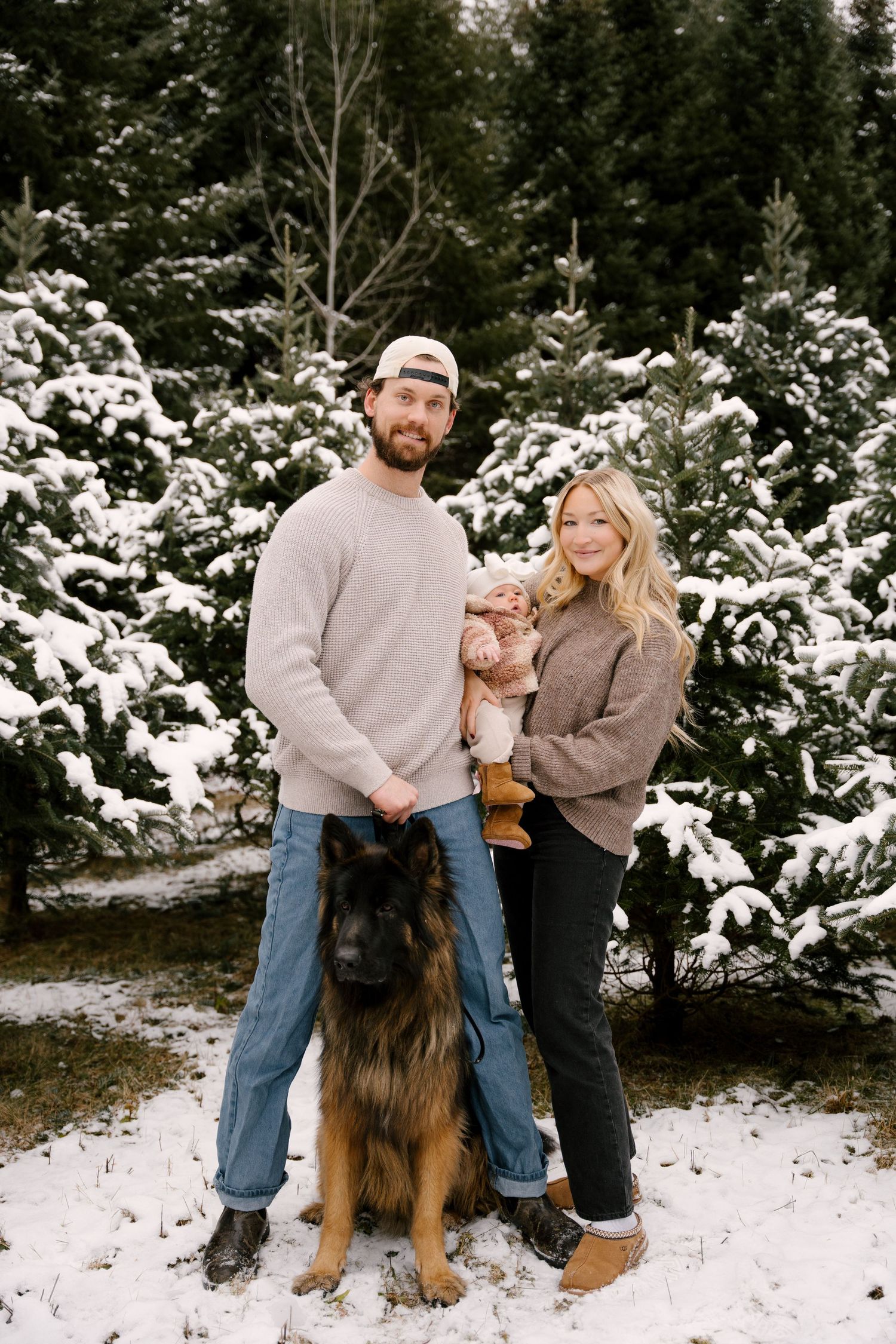 Family portrait at snowy Christmas tree farm with couple holding baby and large German Shepherd dog.