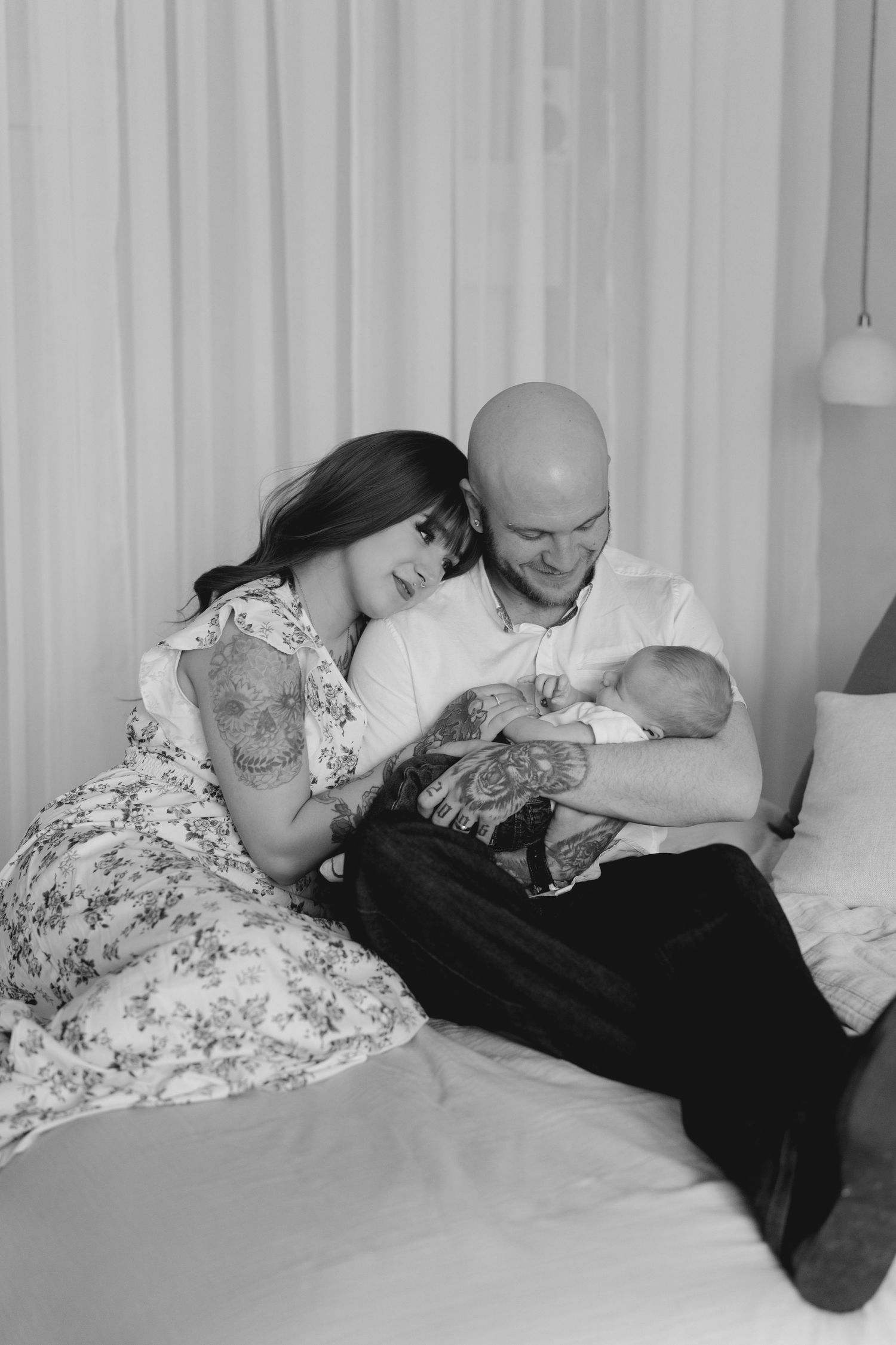 Black and white photo of tattooed parents sitting on bed together smiling while holding their newborn.