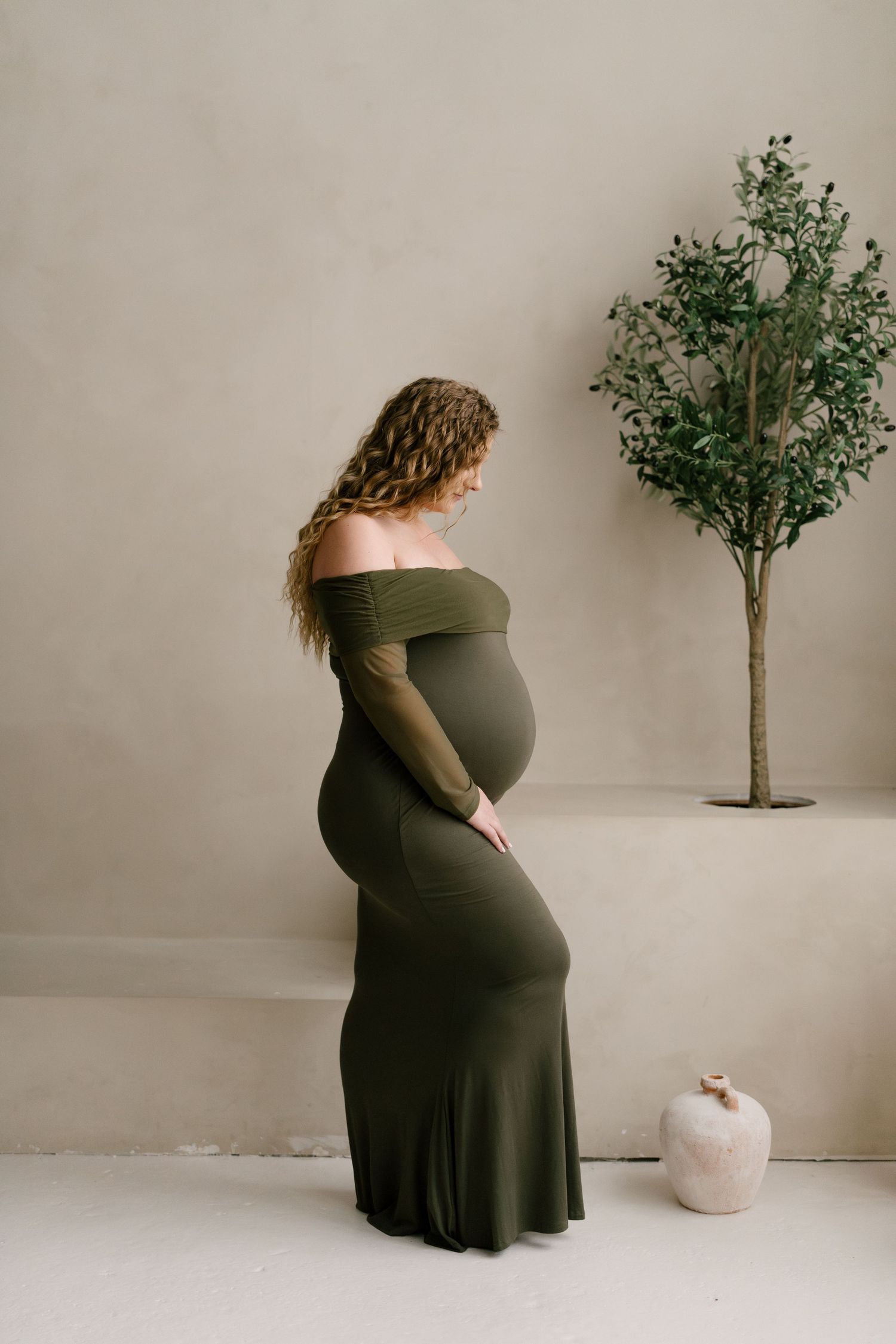 Pregnant woman in olive off-shoulder maxi dress posing beside small indoor tree.