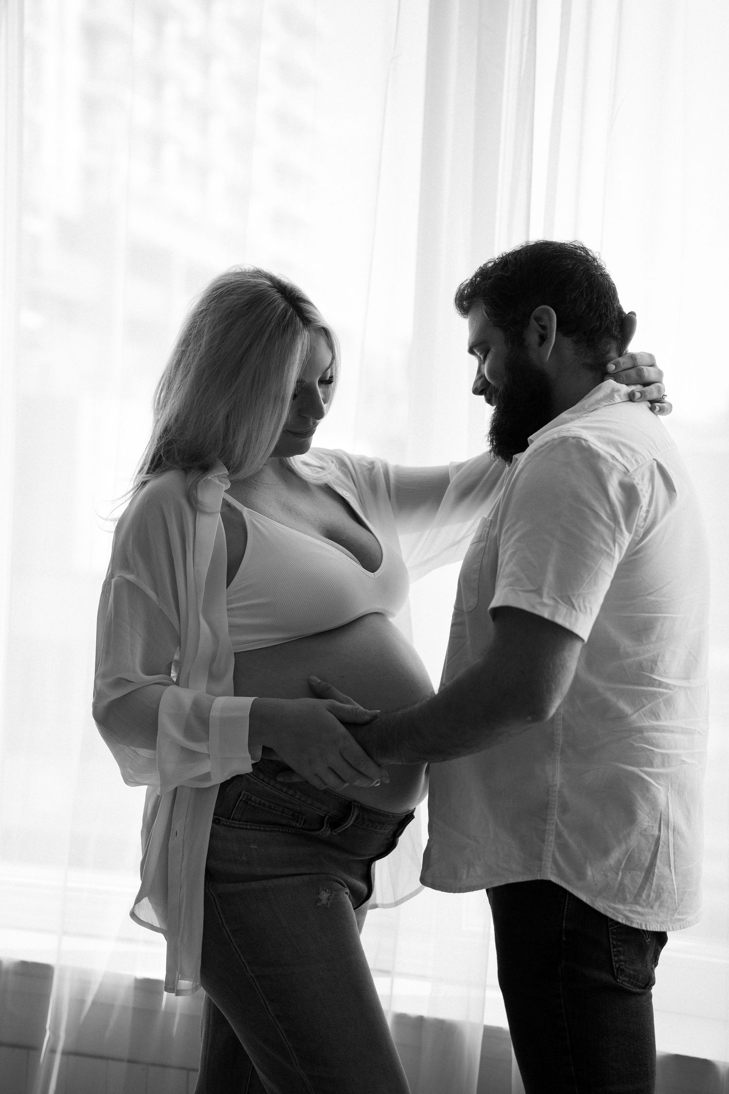 Black and white photo of a couple tenderly cradling a pregnant belly near a bright window.