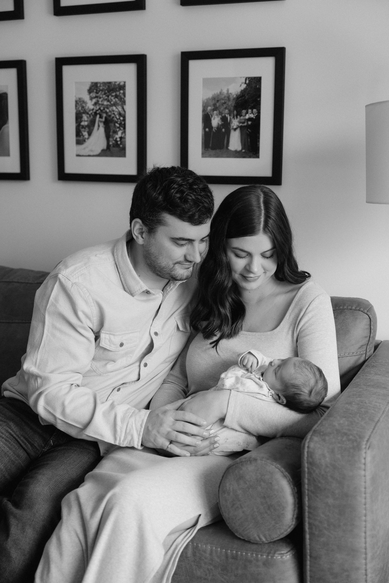 Black and white photo of new parents sitting on a couch, lovingly gazing at their sleeping newborn baby.
