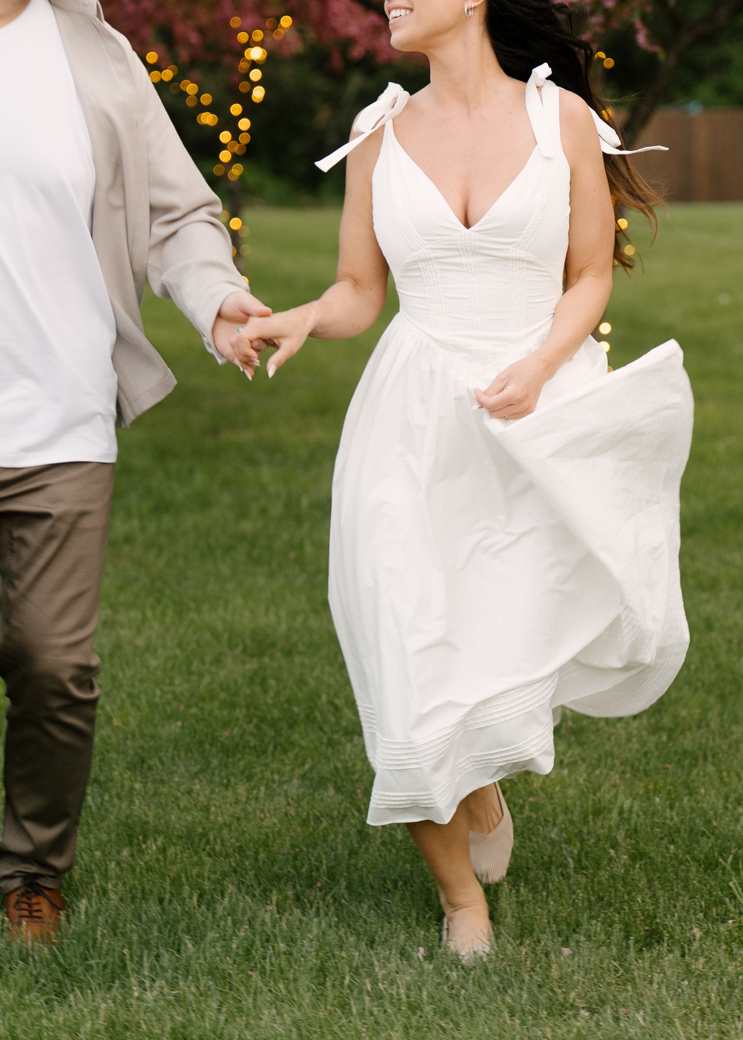 Woman in white sundress with shoulder ties twirling while holding hands with partner on green grass.