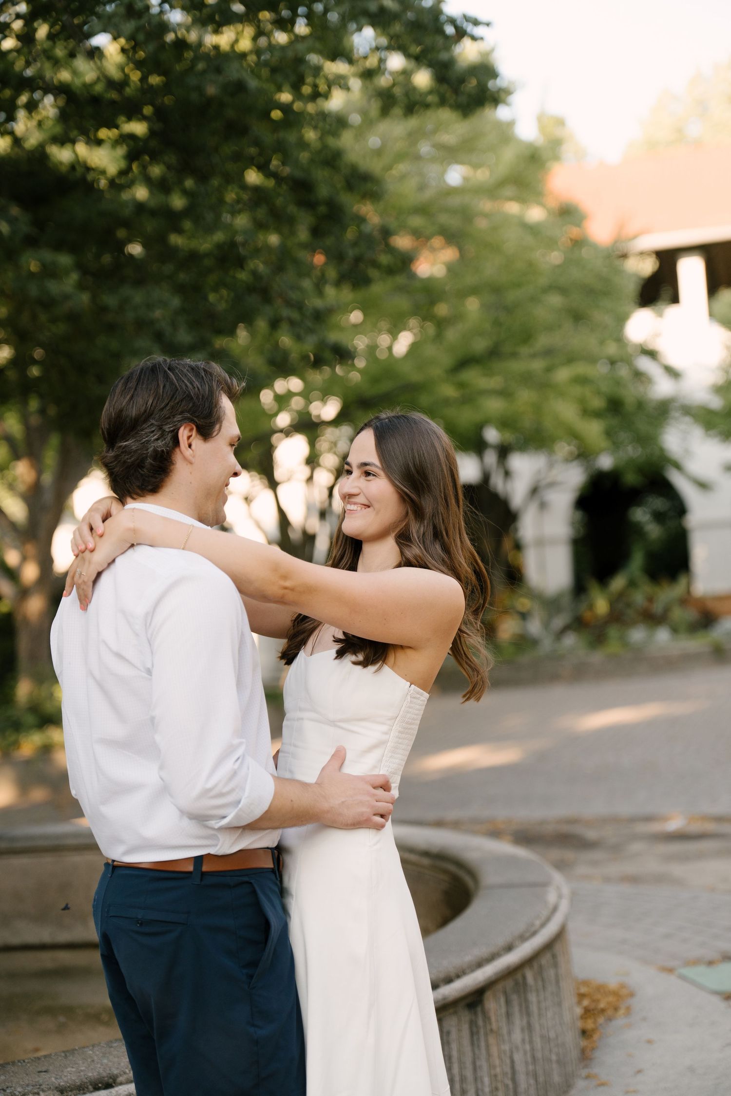 Couple embracing and smiling at each other near a stone fountain in a park.