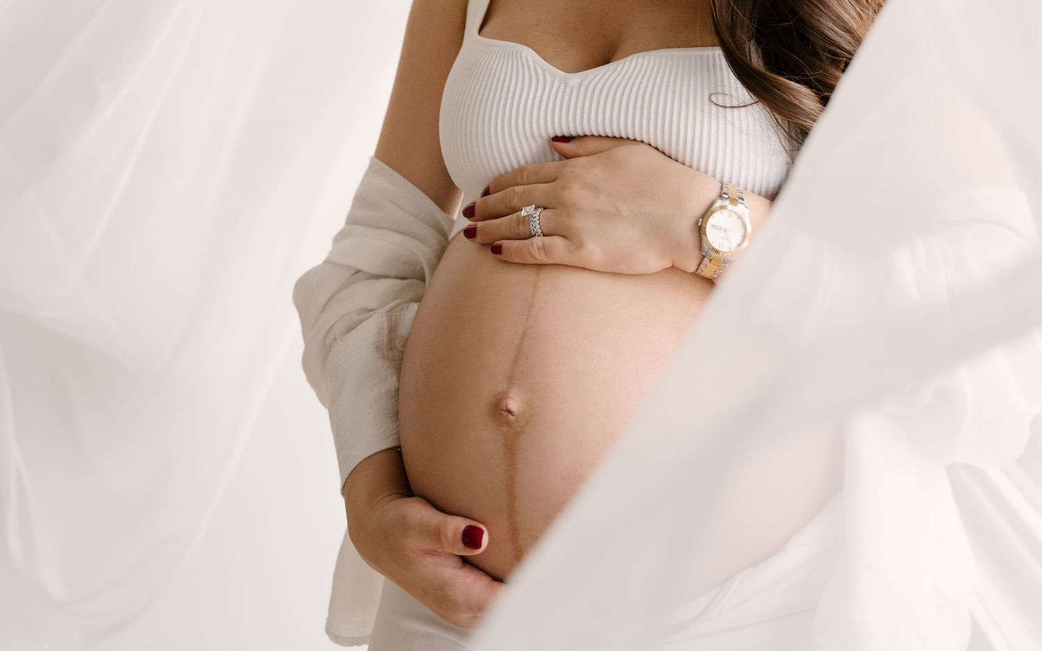 Artistic close-up of pregnant belly with woman in white outfit holding bump, soft flowing fabric in background.