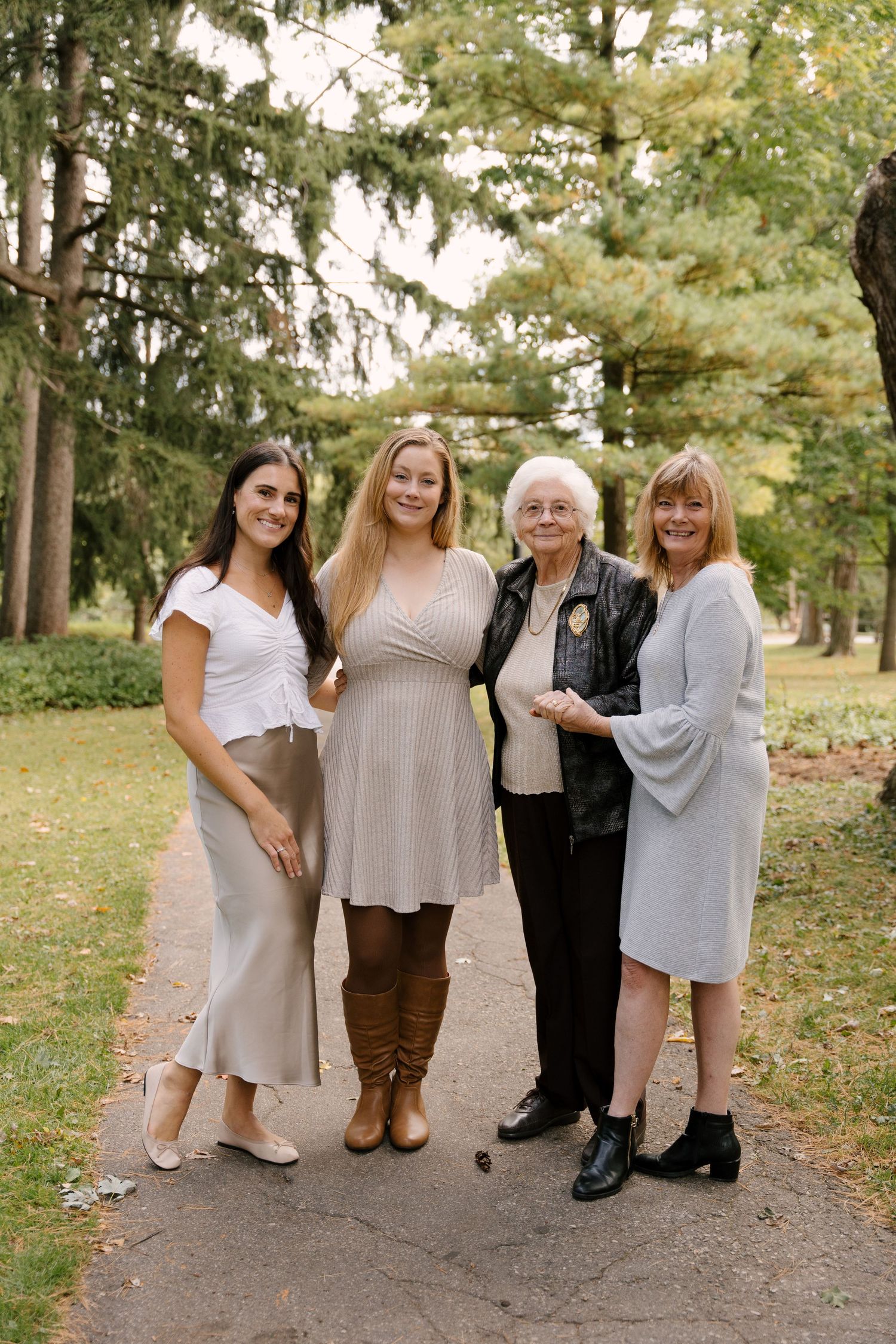 Four women of different generations smiling together on a park pathway surrounded by trees.