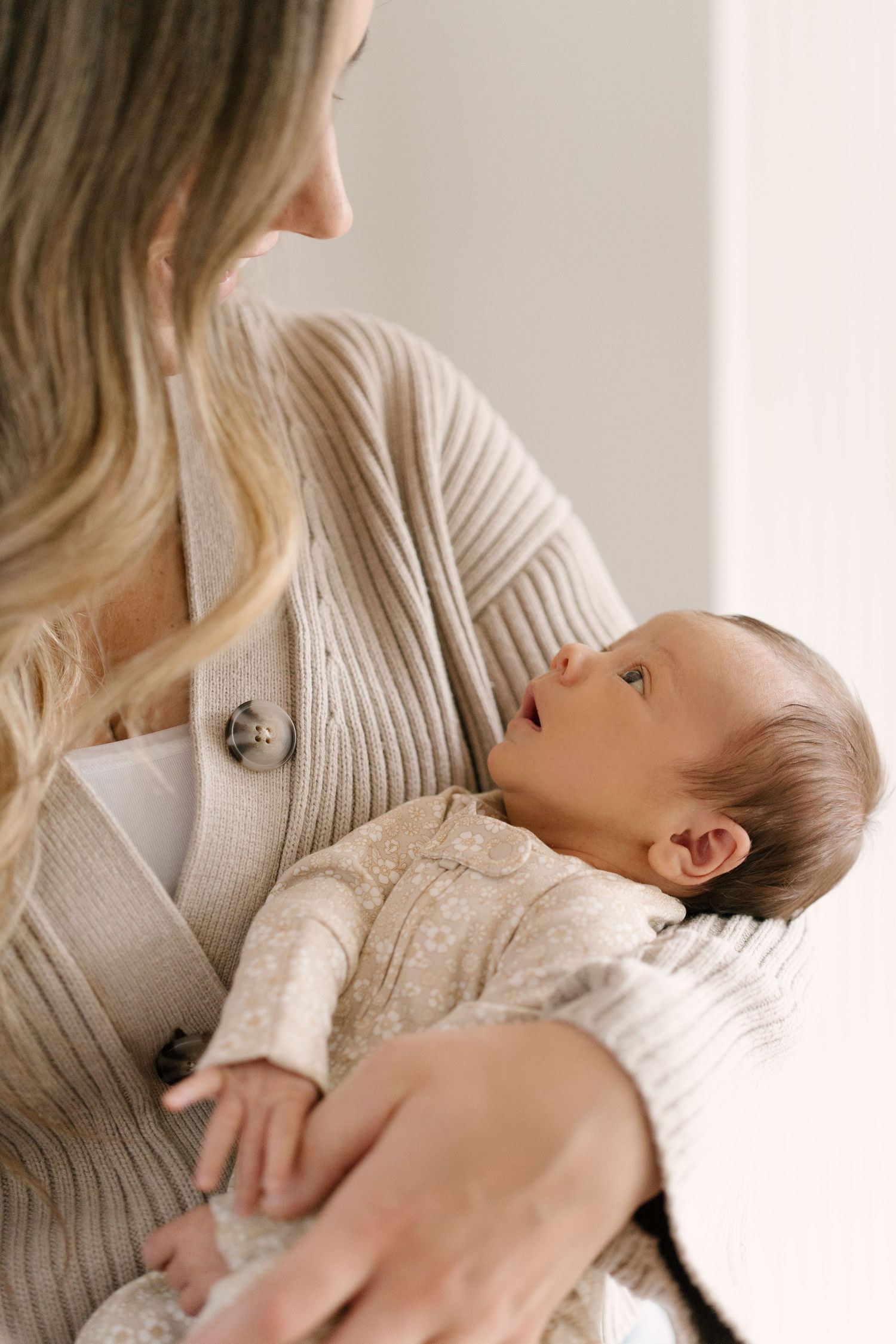A mother in a beige knit cardigan cradles her newborn baby, who gazes up at her adoringly.