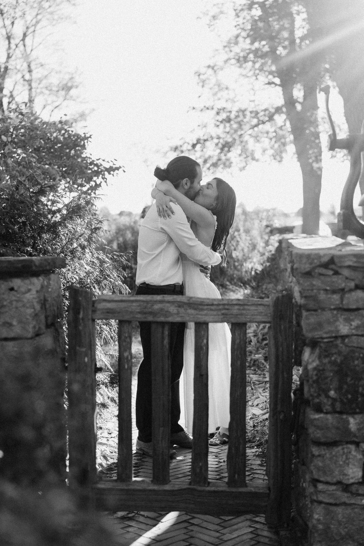 Black and white photo of a couple sharing a romantic kiss by a rustic wooden gate surrounded by lush greenery at Scottsdale Farms.