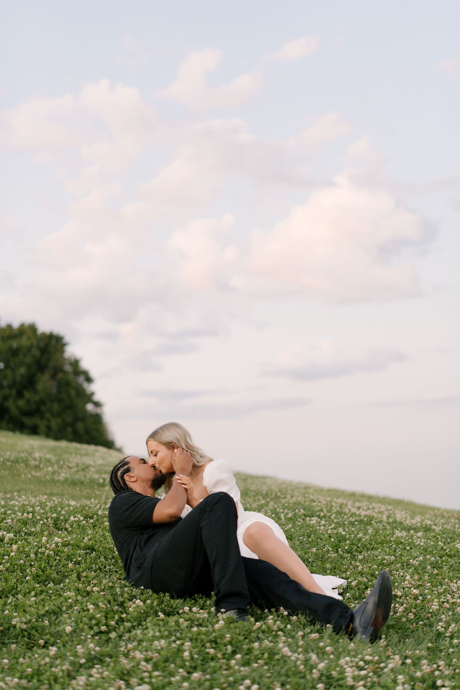 A couple shares a romantic kiss while sitting together on a grassy field with wildflowers under a cloudy sky.
