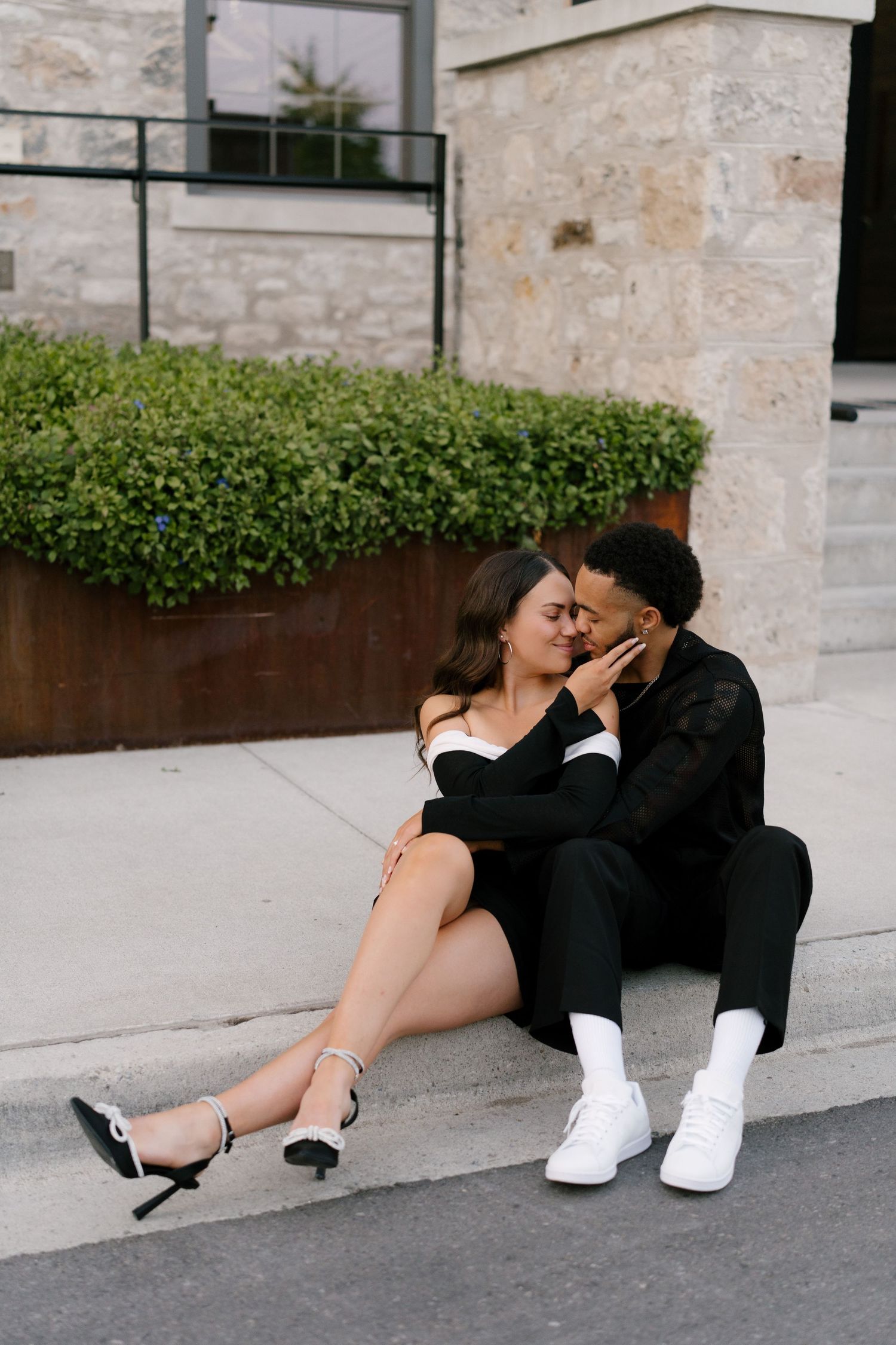 Romantic couple sitting on a curb sharing a tender kiss during an outdoor engagement session.