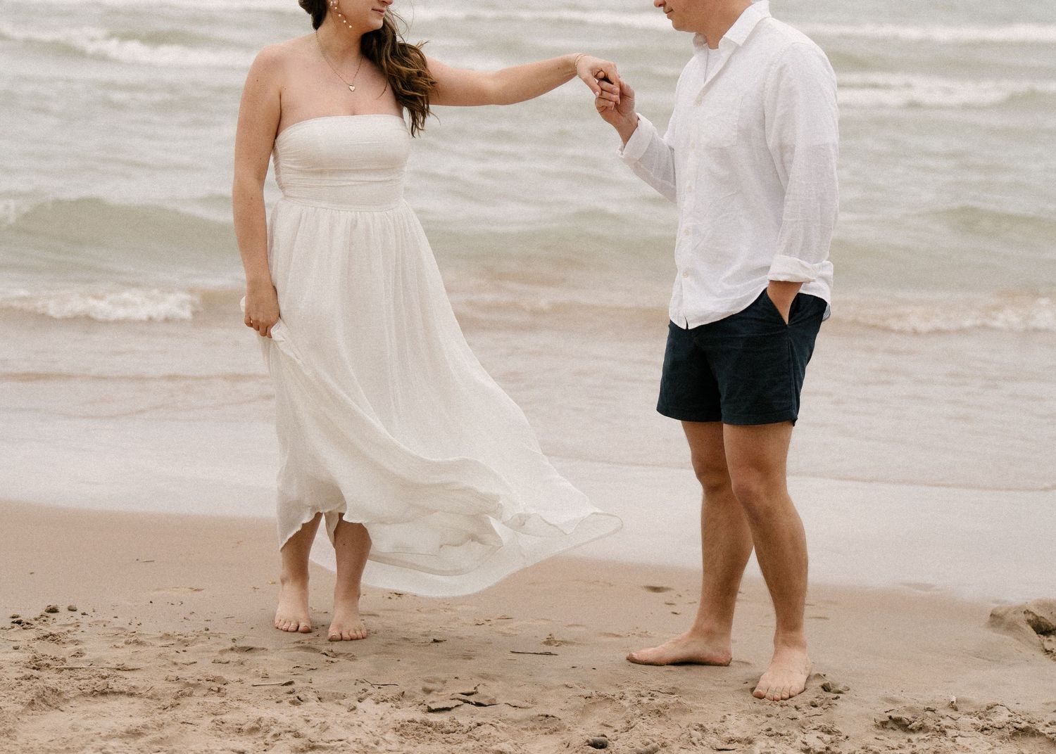 Barefoot couple dancing on beach shore, woman in flowing white strapless dress, man in white shirt and dark shorts.