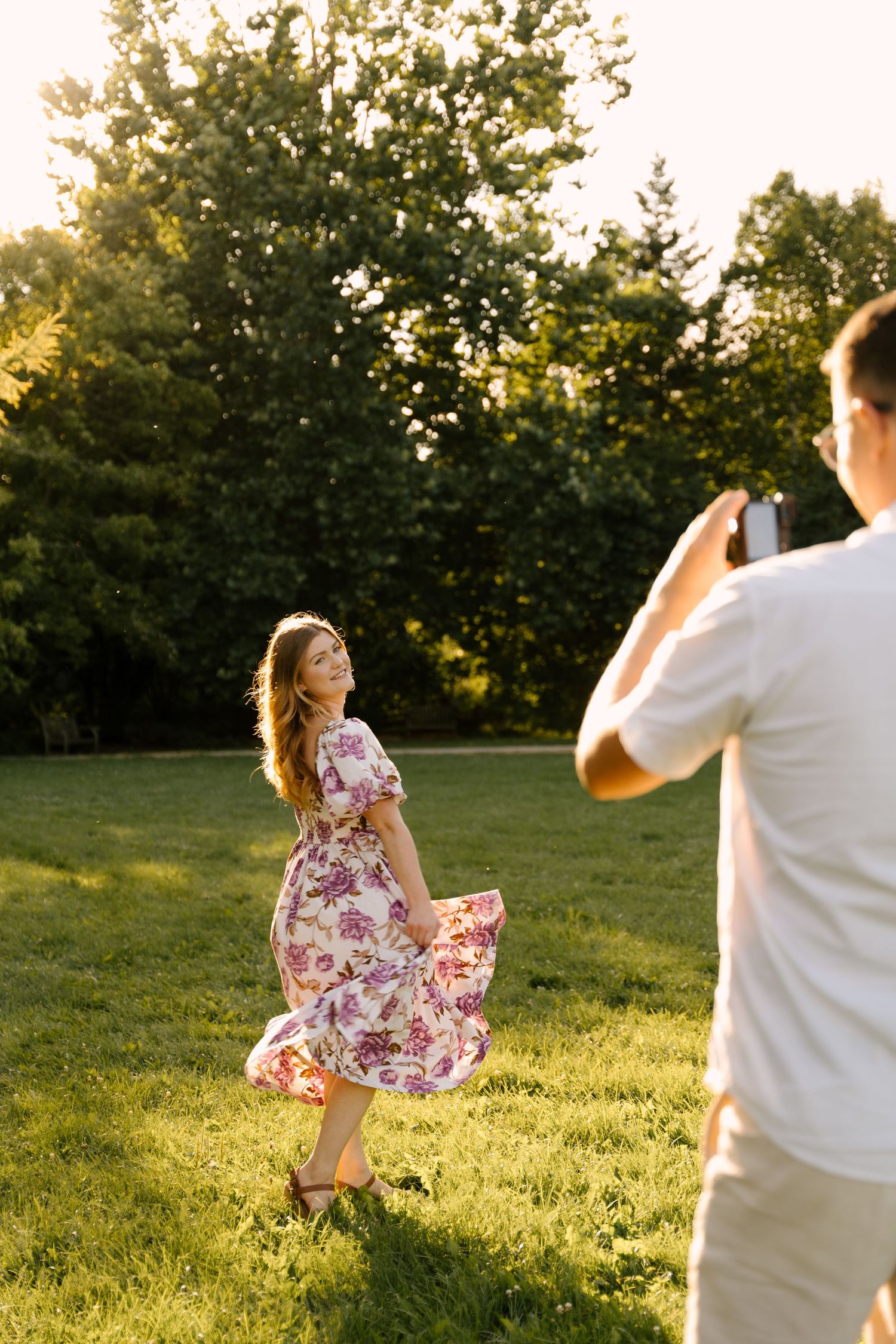 Couple in park at sunset, man photographing woman in floral dress spinning on lush green lawn.