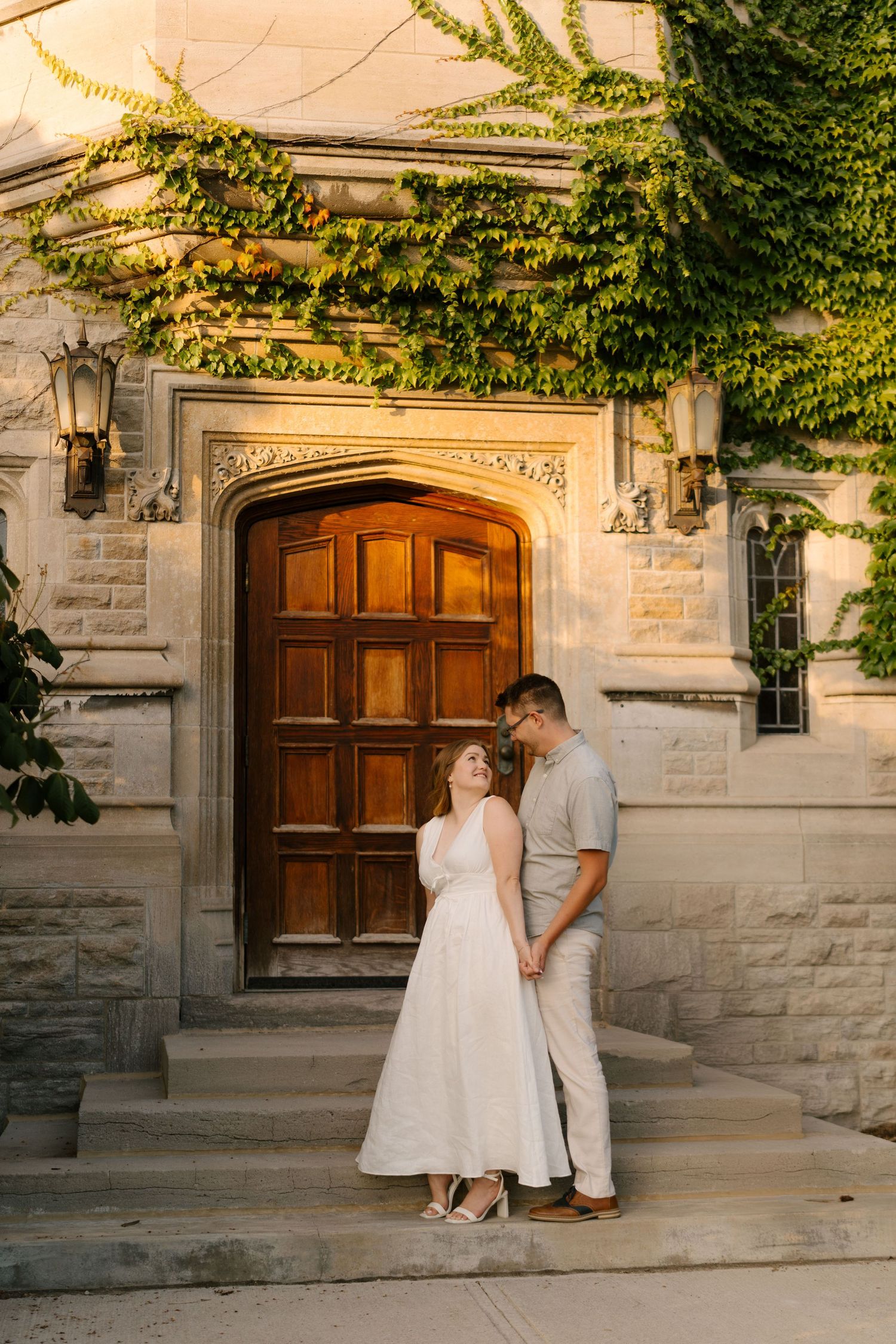Couple holding hands and gazing at each other before ornate wooden door with ivy-covered stone building.