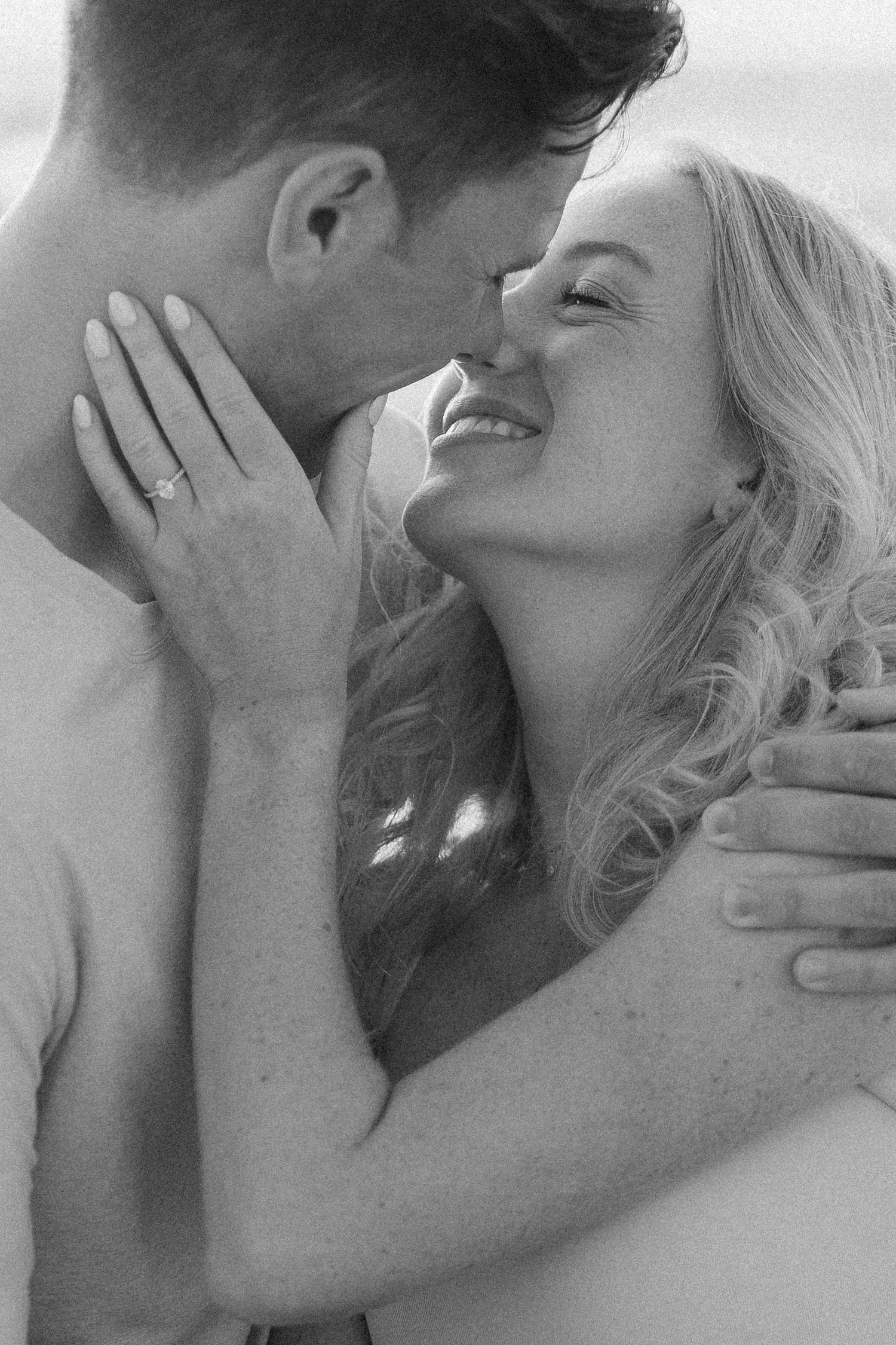 Black and white close-up of smiling woman with engagement ring as partner kisses her cheek.