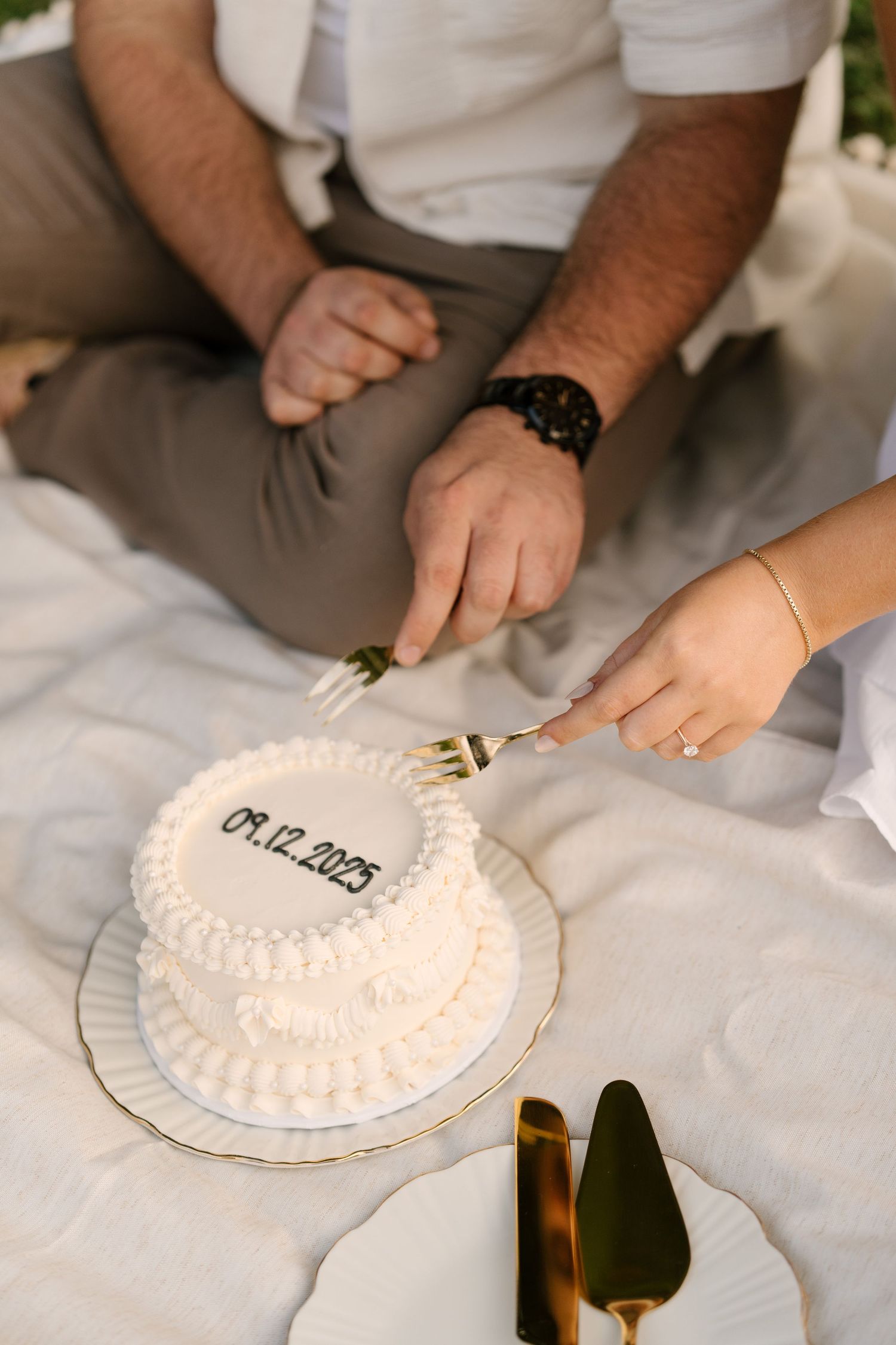 Couple cutting a white two-tier wedding cake dated 09.12.2025 on a picnic blanket outdoors.