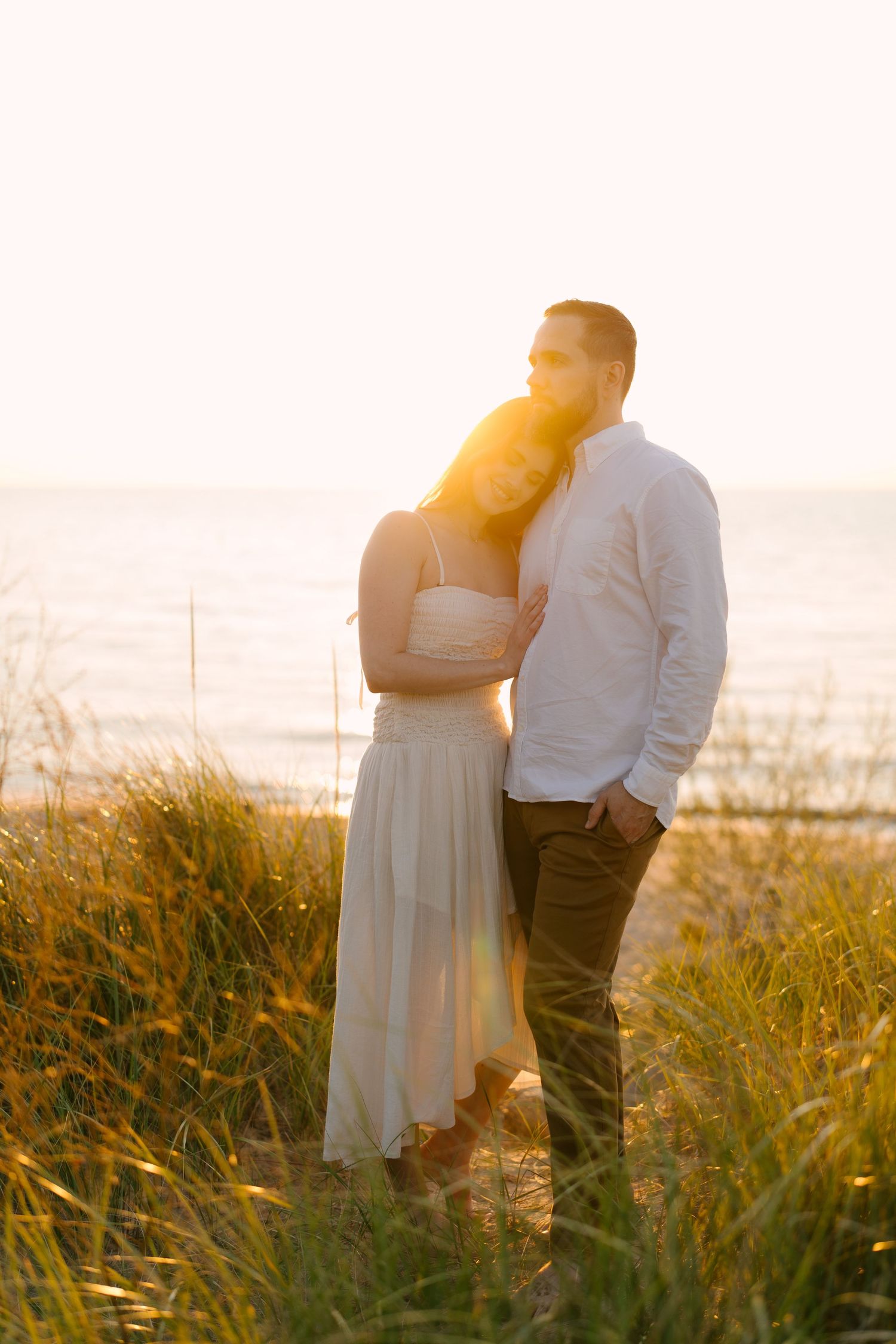 Couple embracing in beach grass at sunset, woman in white dress leaning on man as warm golden light glows behind.