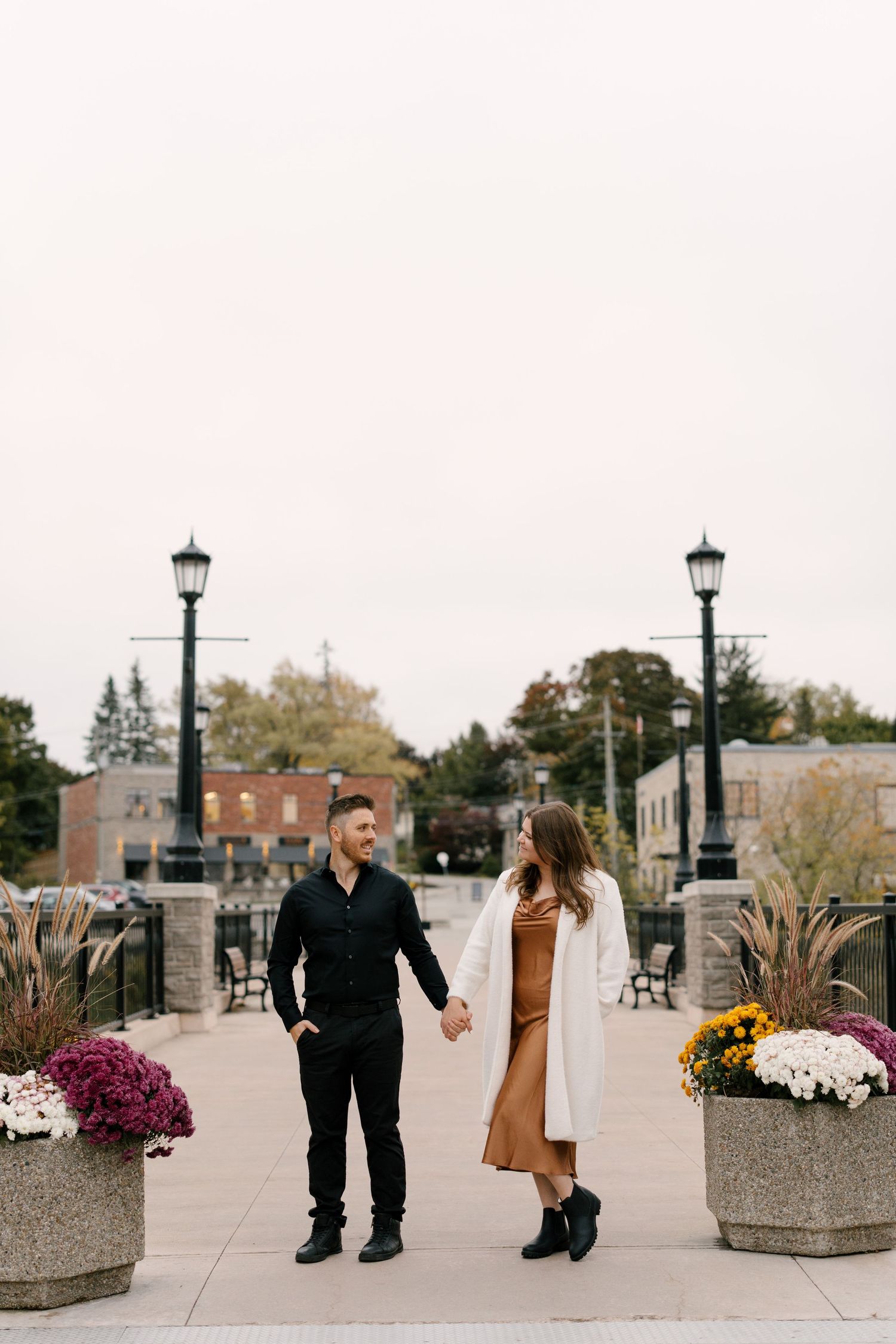 Couple holding hands and gazing at each other on a scenic autumn promenade with colorful mums and lamp posts.