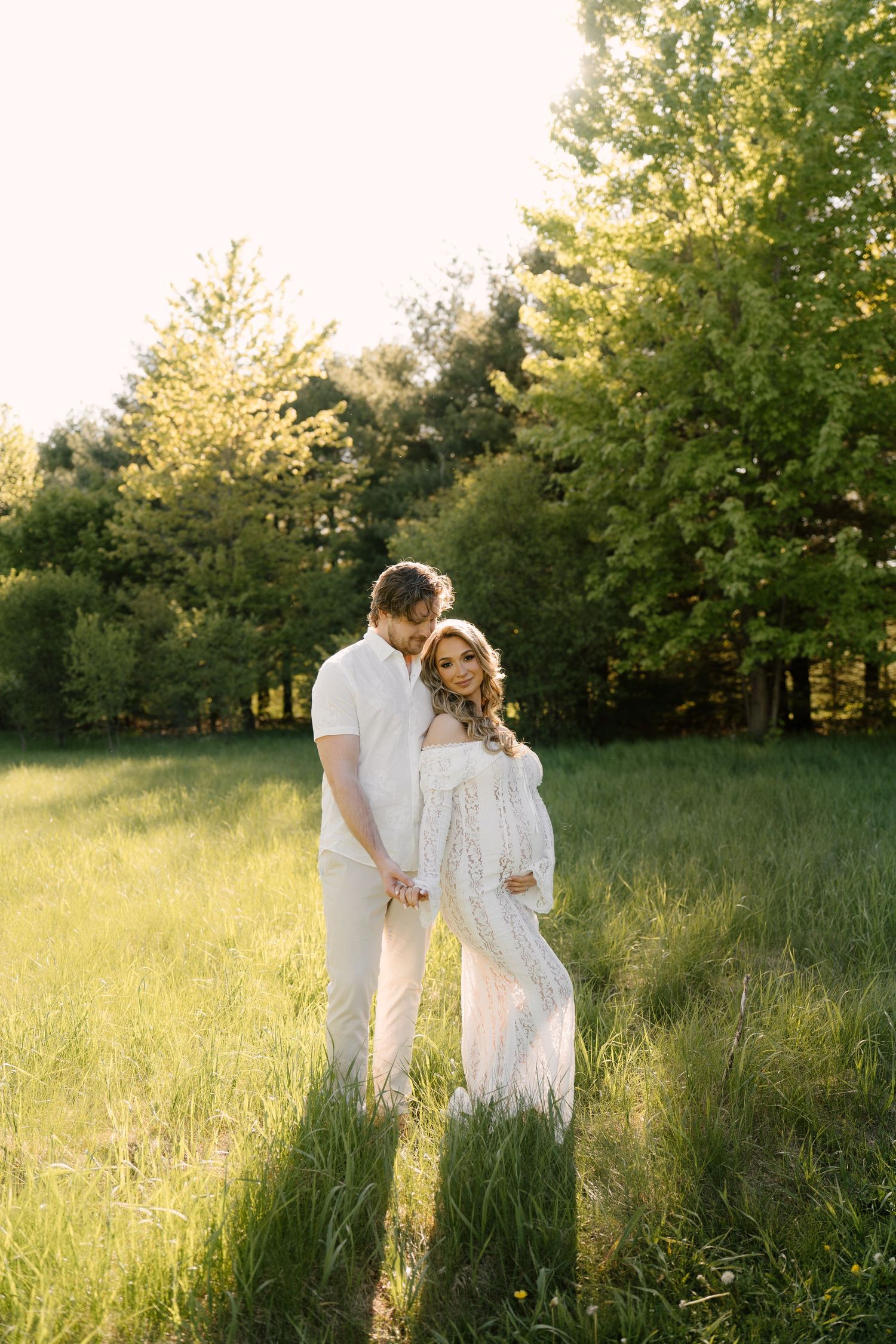 Couple standing close together holding hands in a lush green field during golden hour maternity session.