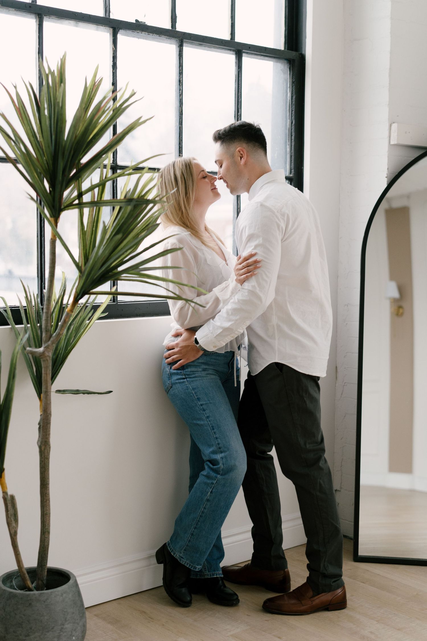 Romantic couple nearly kissing by a large window with a potted plant nearby in a modern space.