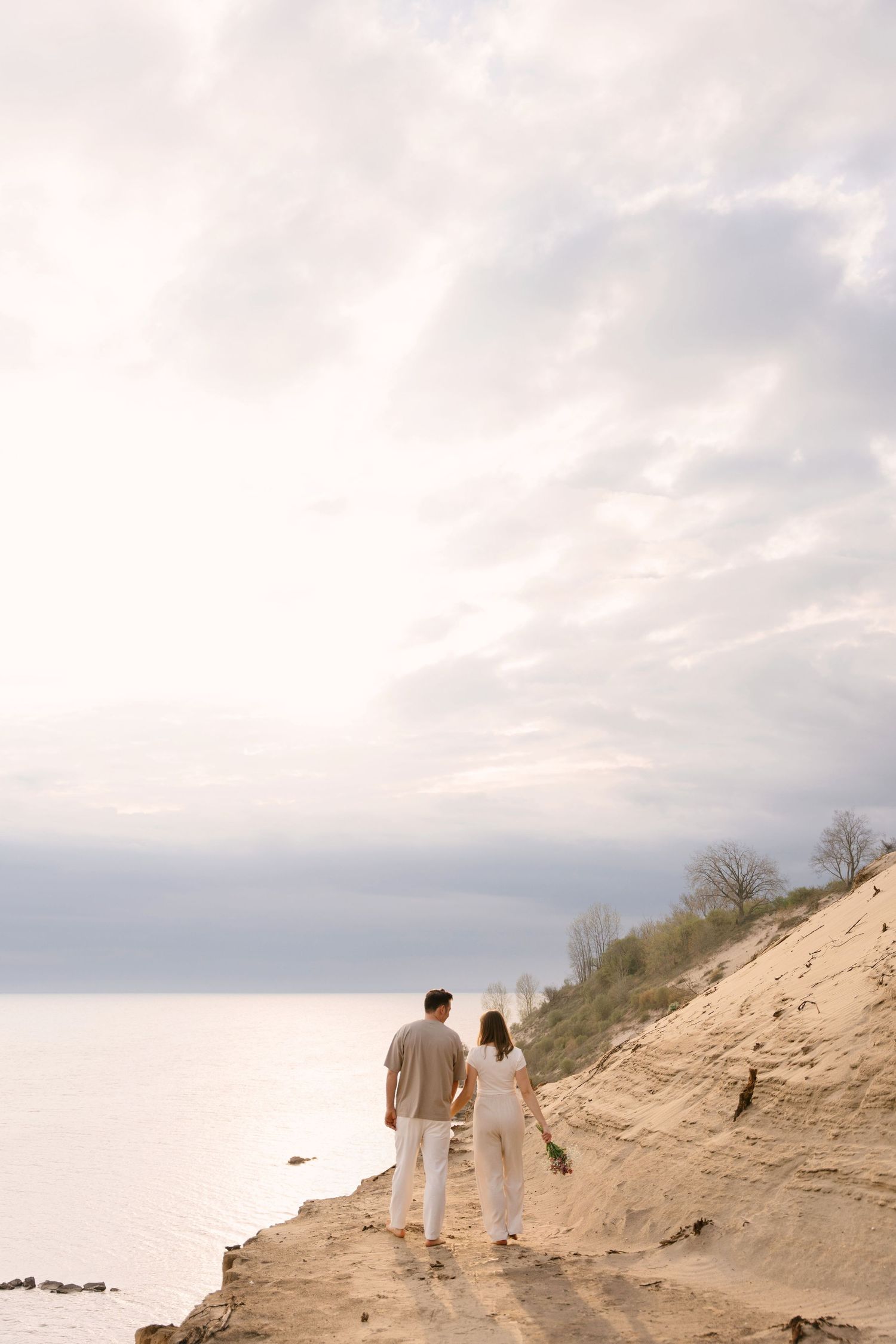 Couple holding hands walking along sandy bluff overlooking calm lake under cloudy sky.