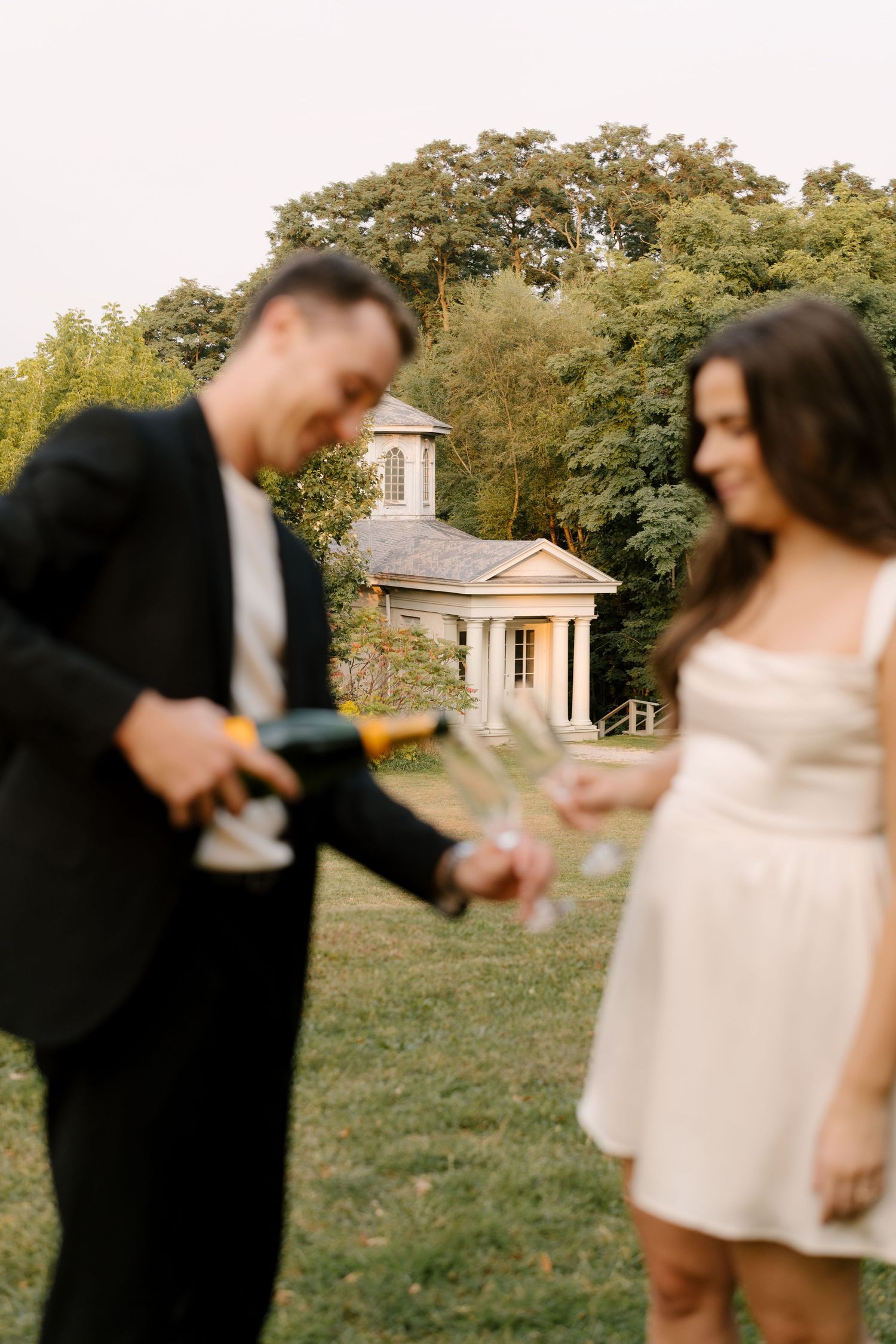 Man pouring champagne into glasses for smiling woman outdoors with classic white gazebo in background.